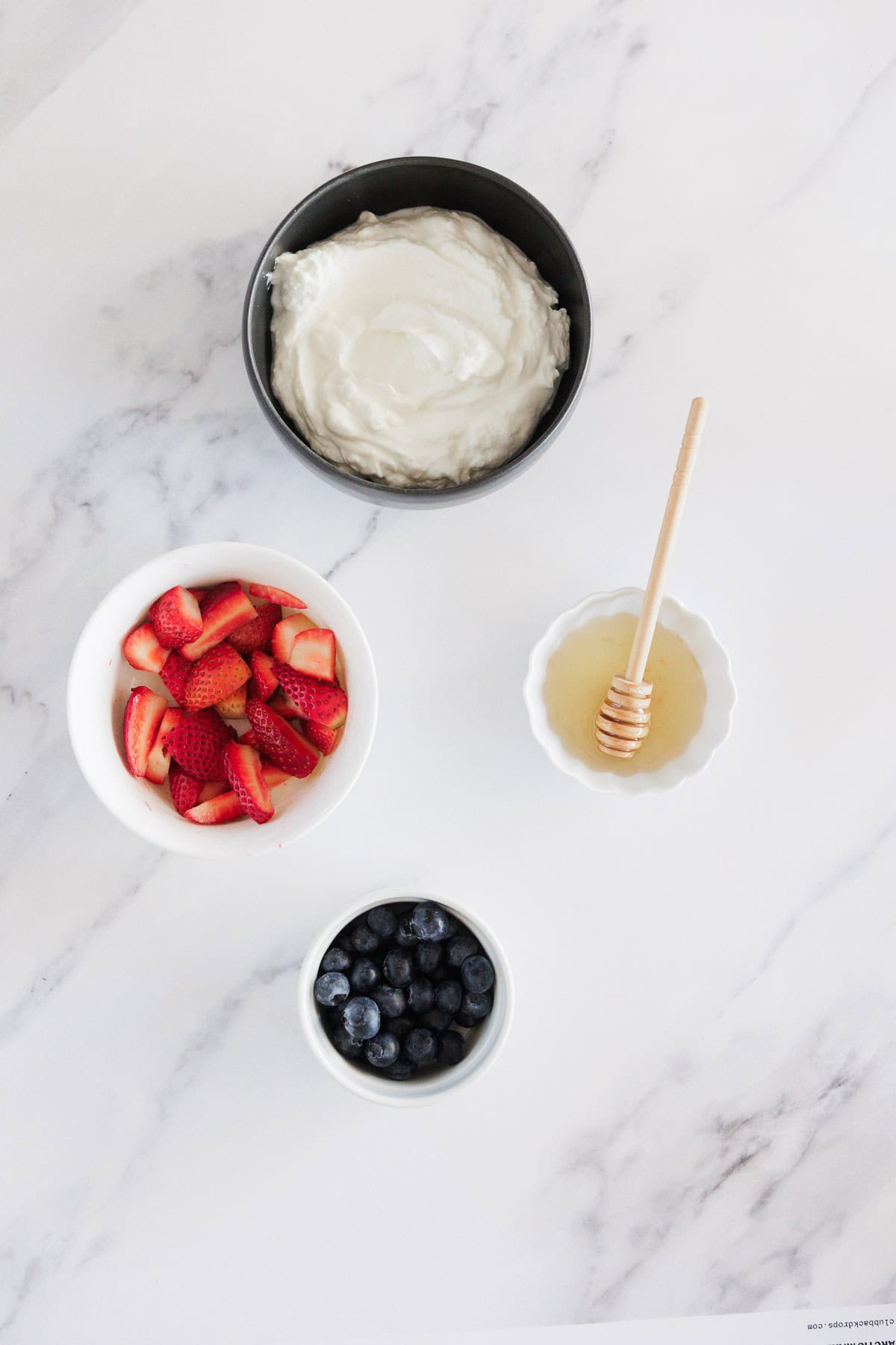 A black bowl of yogurt, a white bowl of sliced strawberries, a white bowl of blueberries, and a small white bowl of honey with a honey dipper, all arranged on a white marble surface.