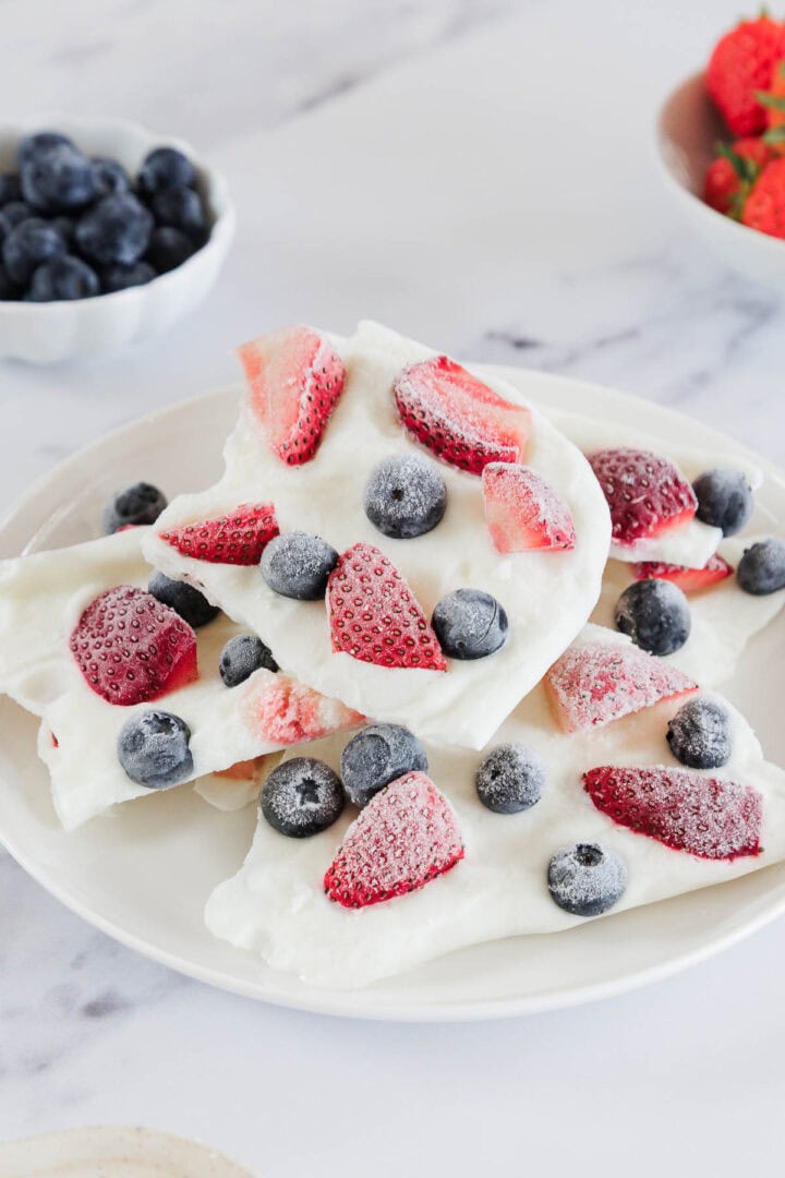A white plate with pieces of yogurt bark topped with frozen strawberries and blueberries. Small bowls of blueberries and strawberries are visible in the background on a white surface.