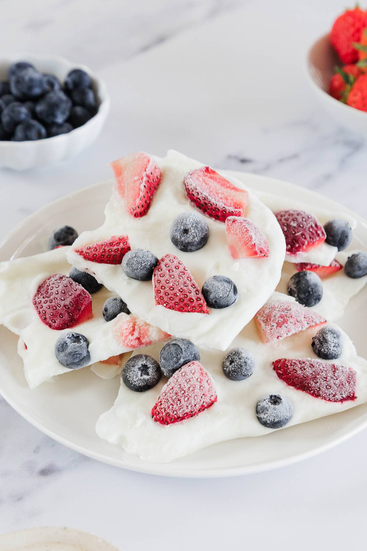 A white plate with pieces of yogurt bark topped with frozen strawberries and blueberries. Small bowls of blueberries and strawberries are visible in the background on a white surface.