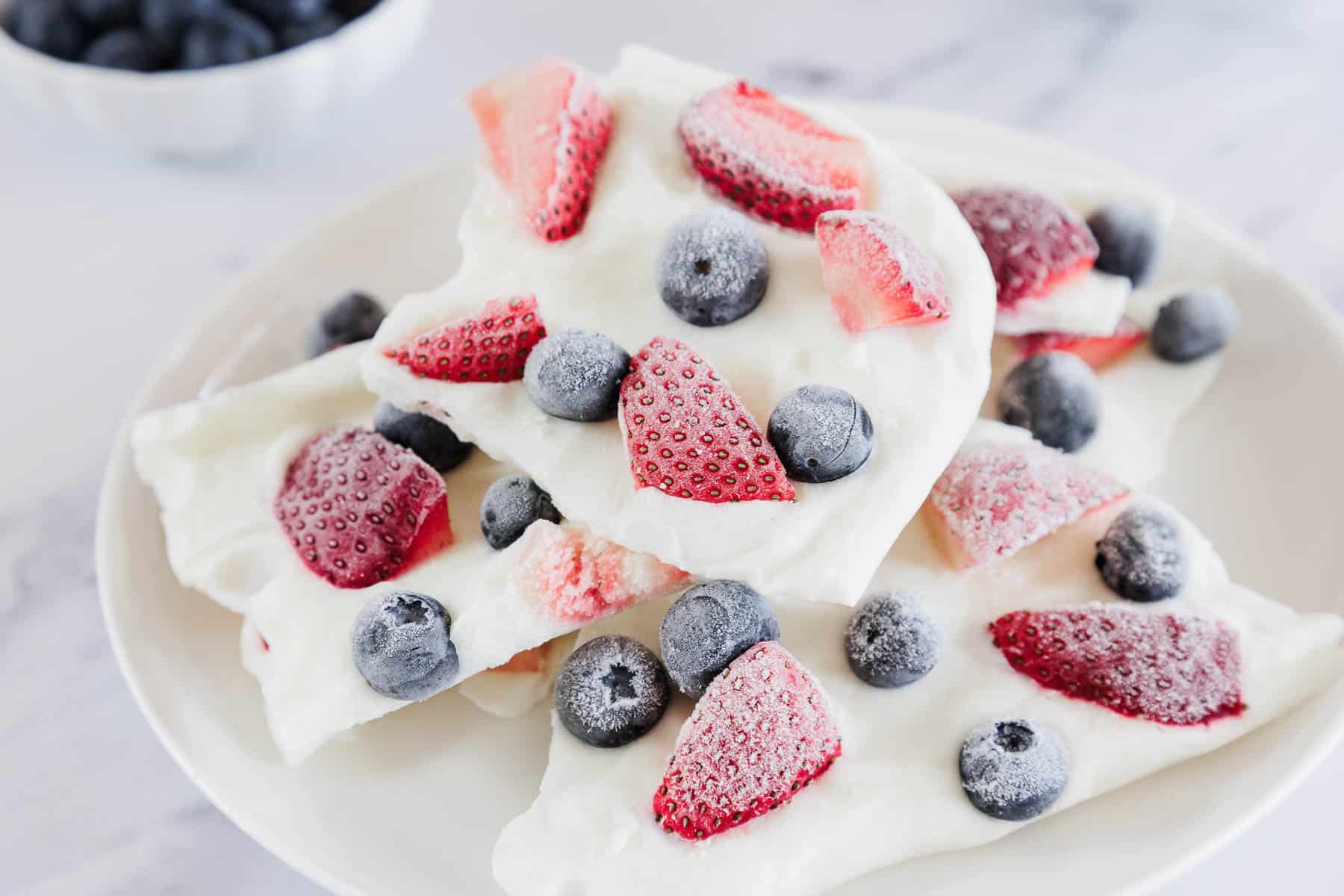 A white plate with pieces of frozen yogurt bark topped with sliced strawberries and whole blueberries. A small bowl of blueberries is in the background.