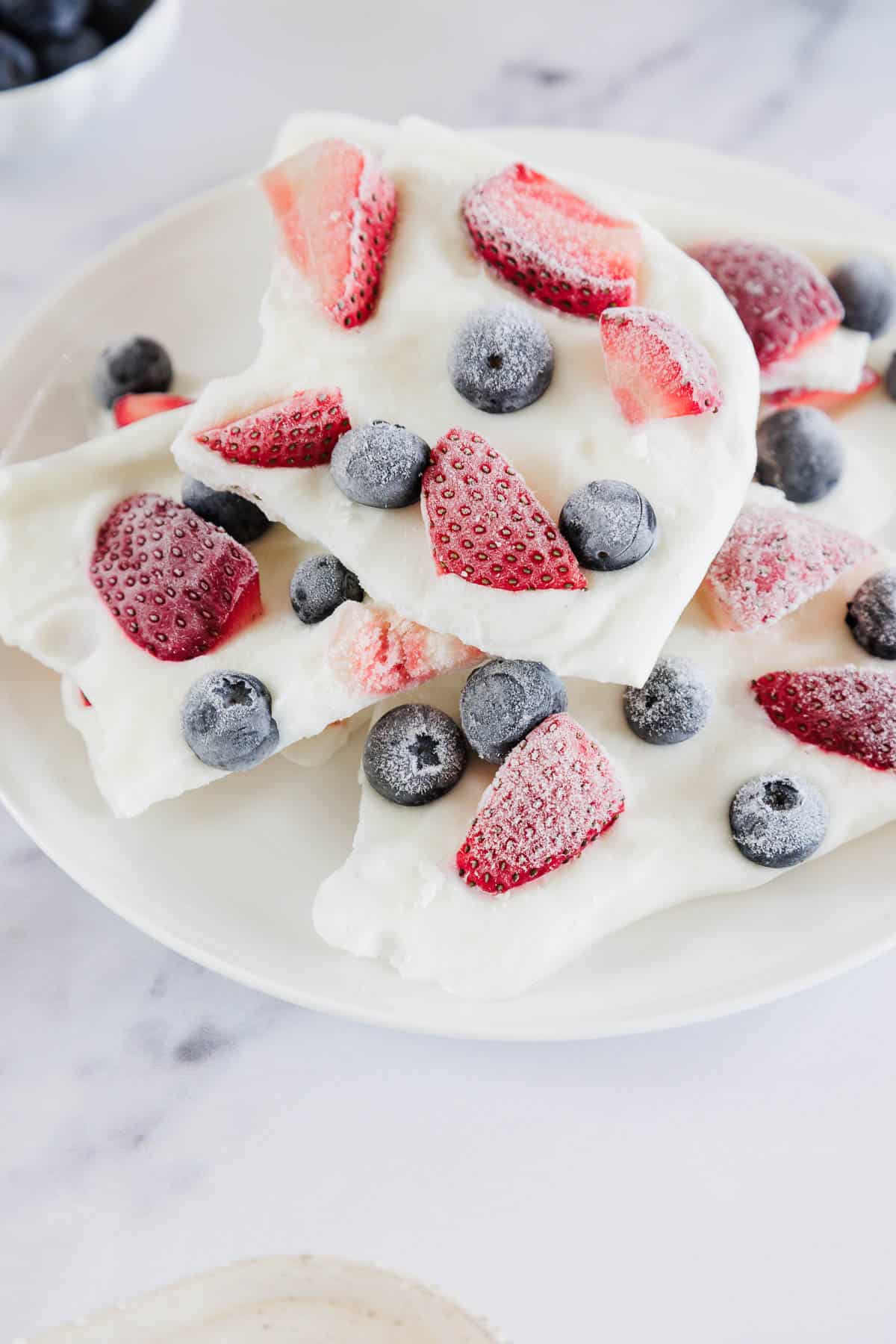 A white plate with pieces of yogurt bark topped with frozen strawberries and blueberries, set on a light marble surface.