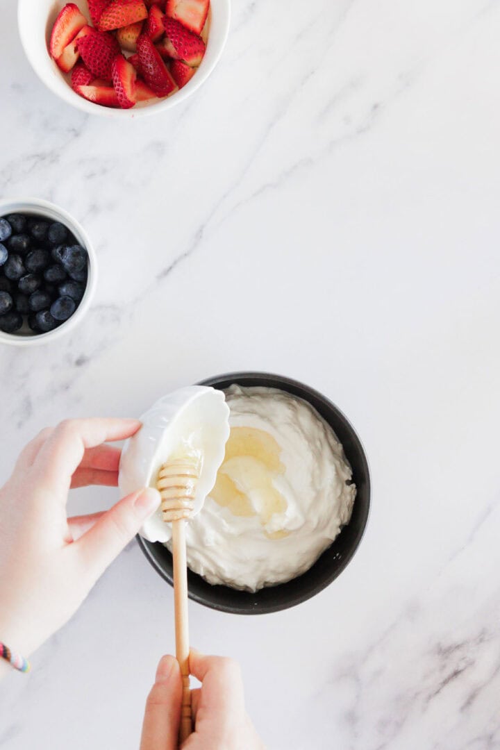 A person drizzles honey into a bowl of yogurt with a honey dipper. Nearby are small bowls containing sliced strawberries and blueberries on a white marble surface.