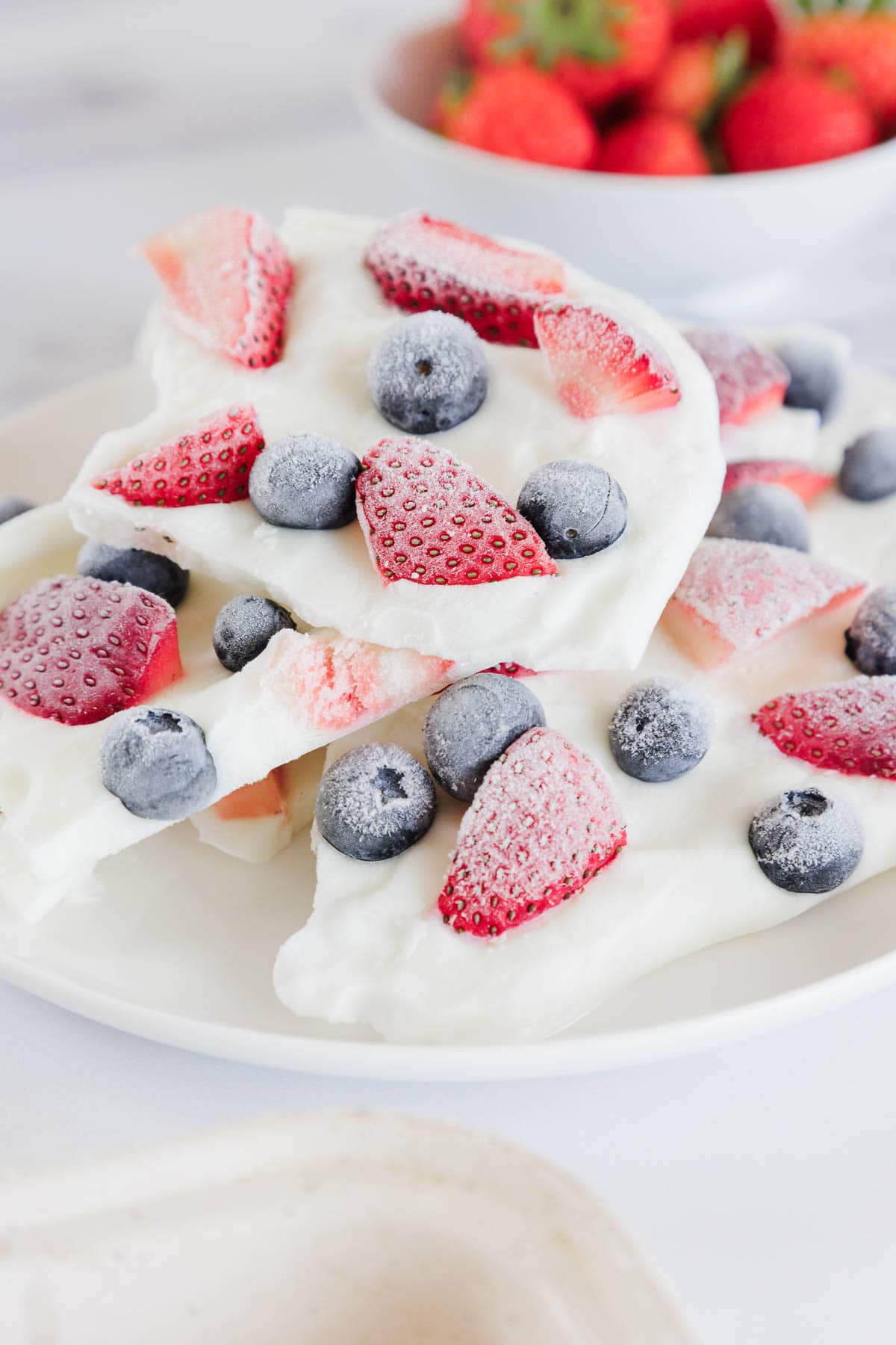 A plate of yogurt bark topped with frozen strawberries and blueberries is shown. A bowl of fresh strawberries is in the background. The yogurt bark pieces are stacked and feature visible fruit on top.