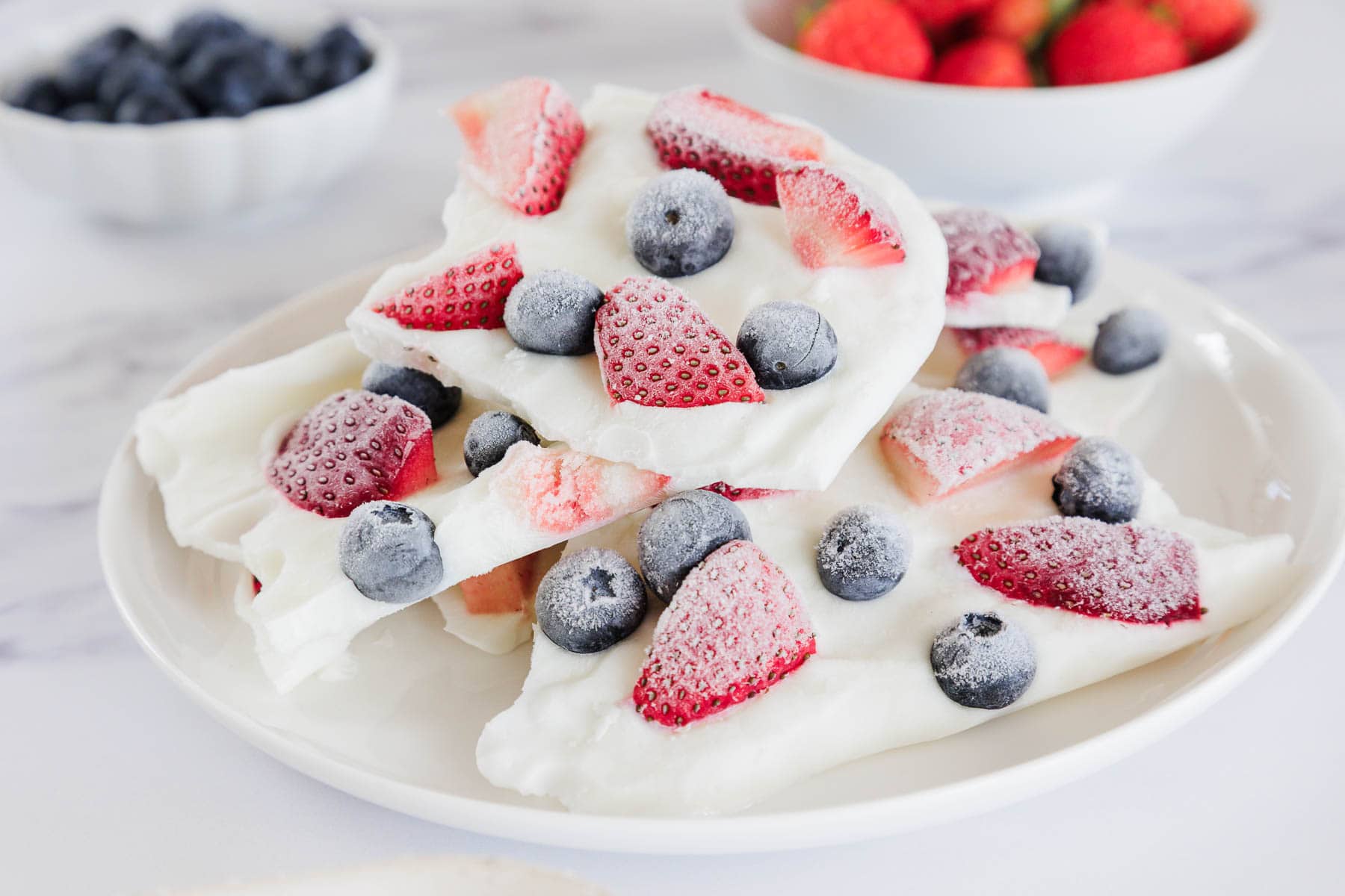 A white plate with pieces of frozen yogurt bark topped with blueberries and sliced strawberries. In the background, there are bowls of whole blueberries and strawberries on a white surface.