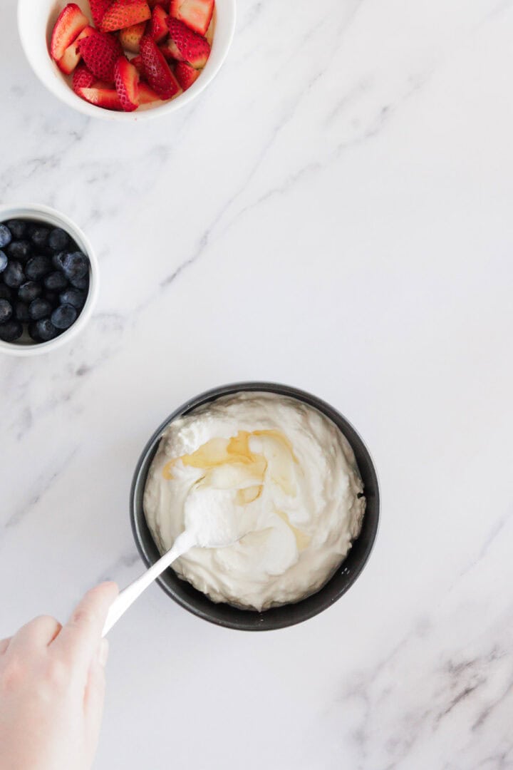 A hand holds a spoon mixing yogurt with honey in a black bowl. Nearby, there is a white bowl of blueberries and a white bowl of sliced strawberries on a white marble surface.