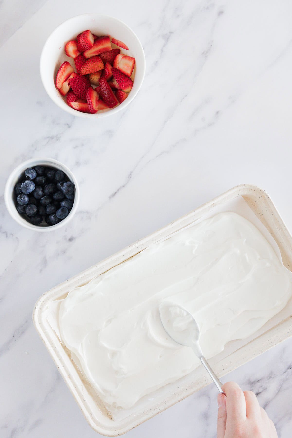 A hand spreads a layer of white creamy mixture in a rectangular dish with a spoon. Nearby, a bowl of sliced strawberries and a bowl of blueberries sit on a white marble surface.
