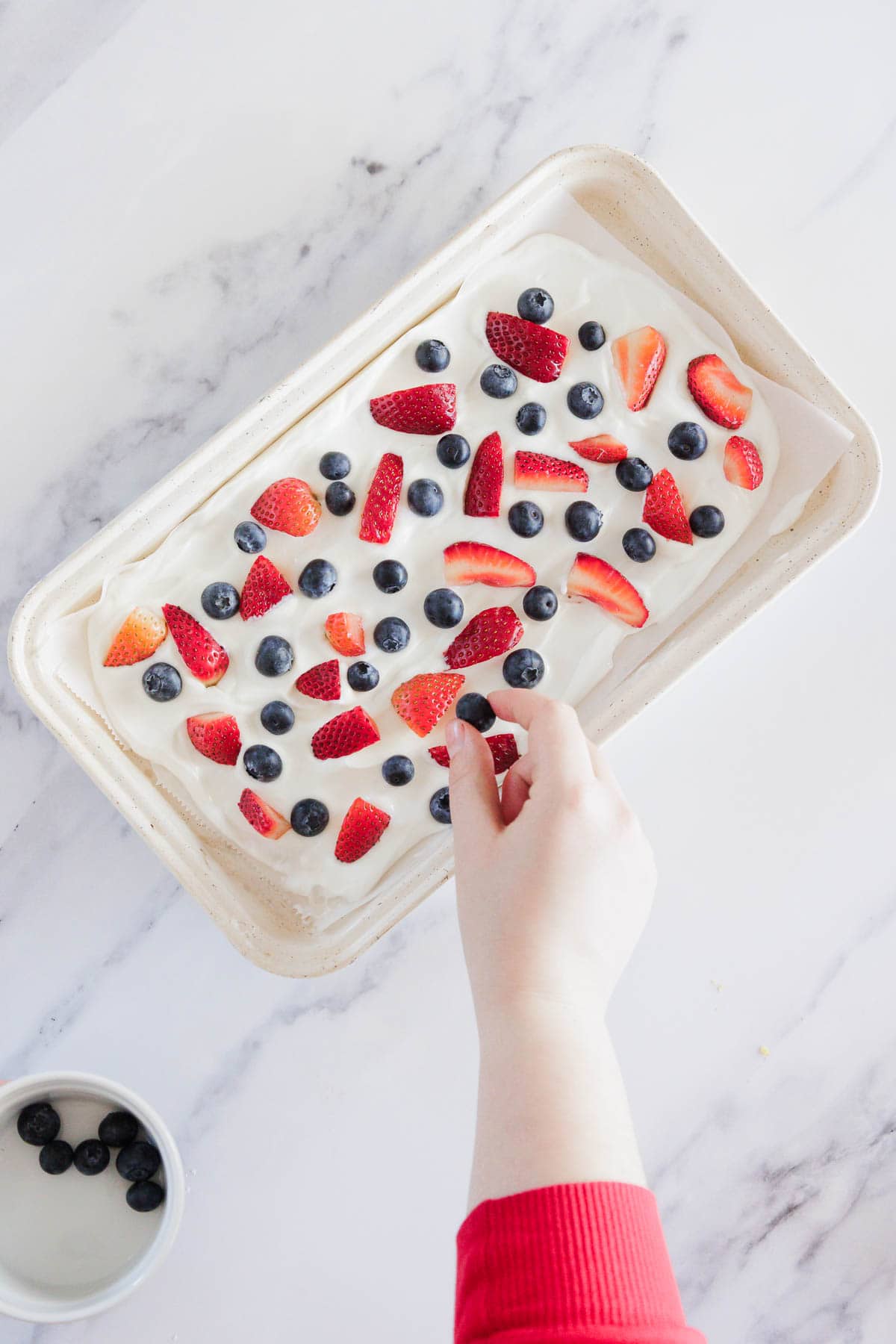 A hand places a blueberry onto a rectangular dish filled with white yogurt and topped with blueberries and sliced strawberries, on a white marble surface. A small bowl with more blueberries is nearby.