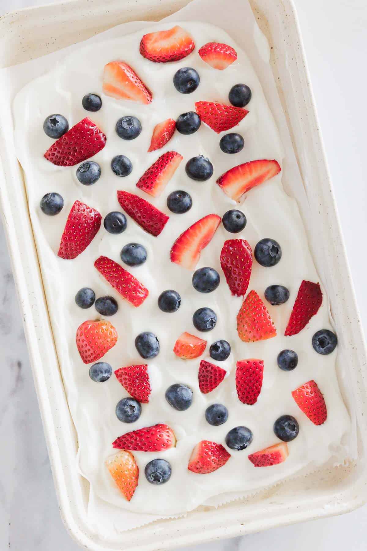 A rectangular pan filled with a layer of white whipped cream or yogurt, topped with whole blueberries and sliced strawberries, arranged evenly across the surface.