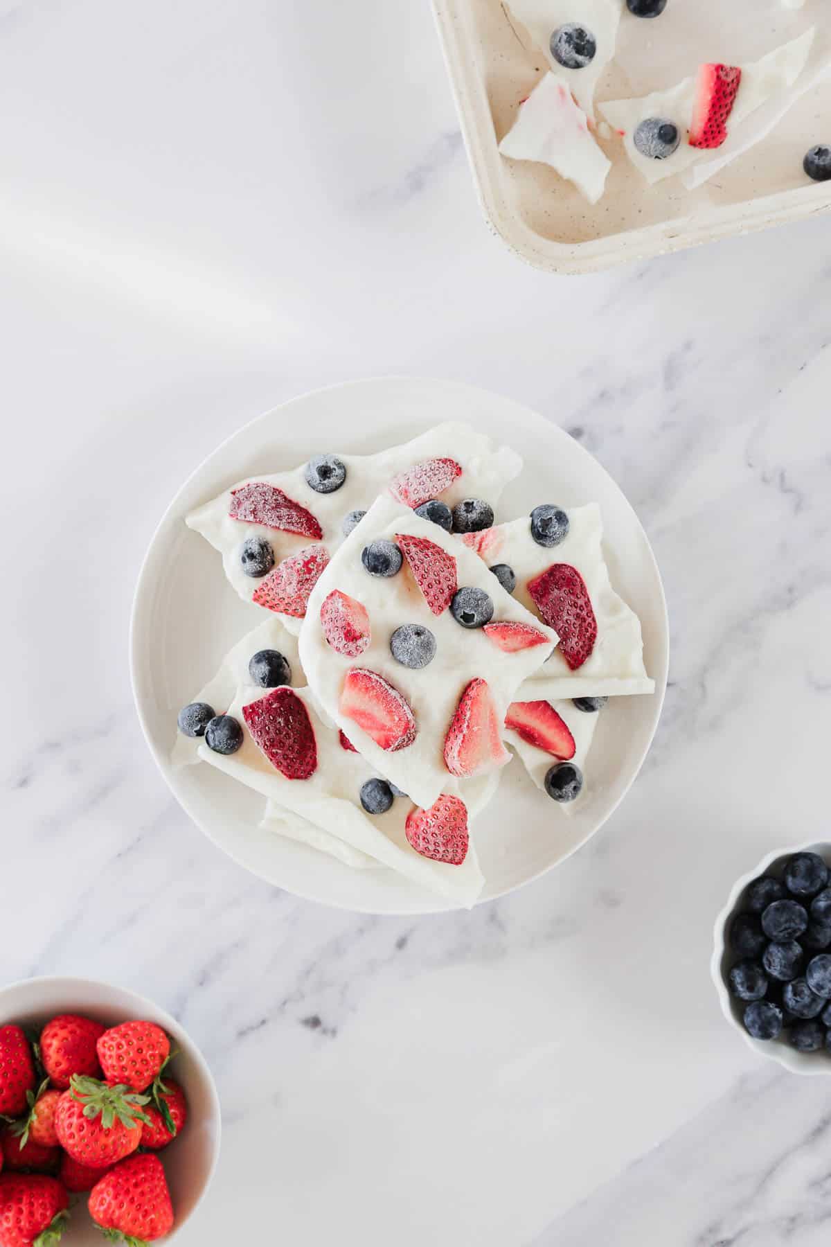A white plate with pieces of yogurt bark topped with strawberries and blueberries. Nearby are a bowl of fresh blueberries, a bowl of strawberries, and a tray with more yogurt bark pieces on a white marble surface.
