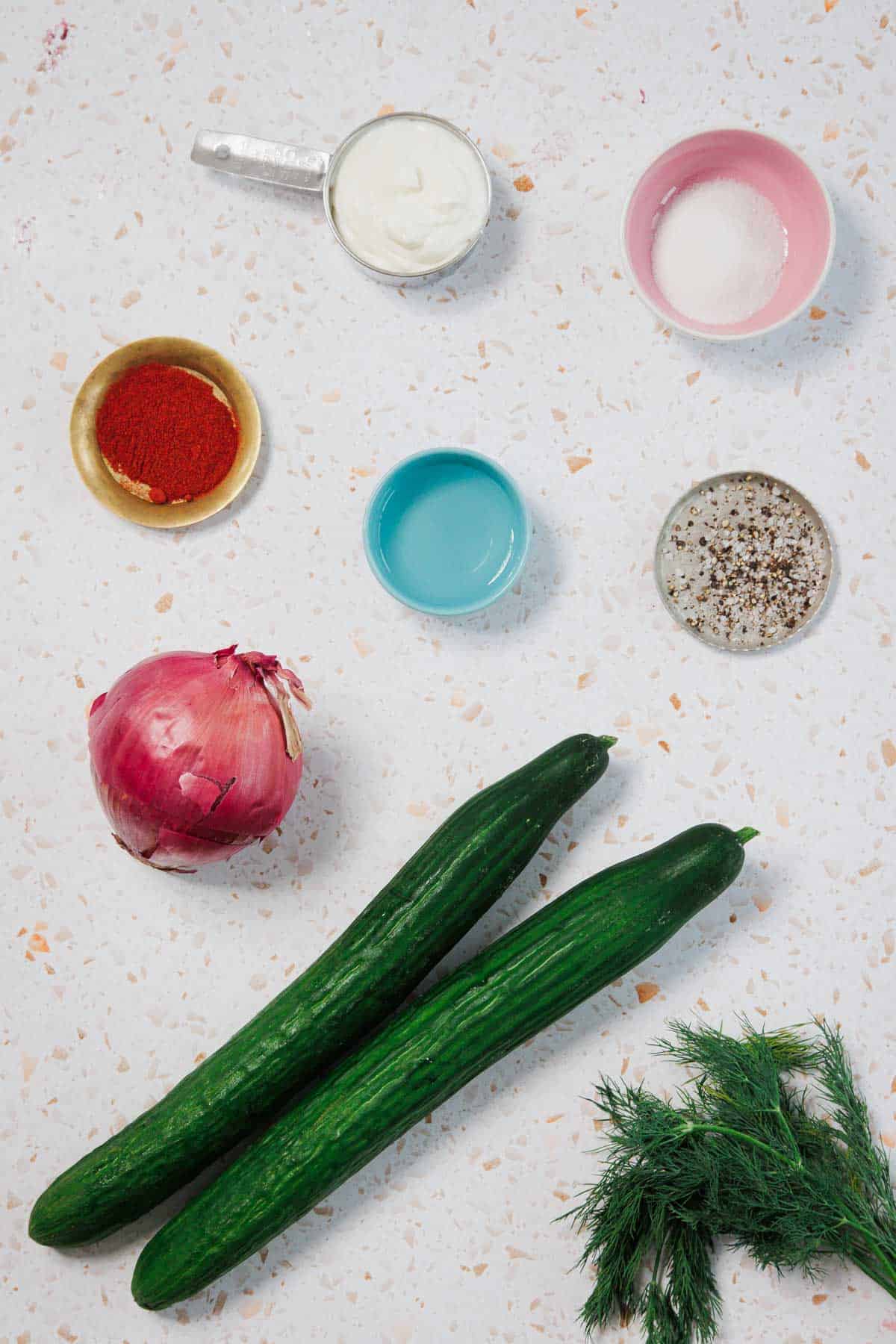 Ingredients on a white speckled surface, including two cucumbers, a bunch of dill, a red onion, paprika, black pepper, salt, a small bowl of white yogurt, and a small bowl of clear liquid.