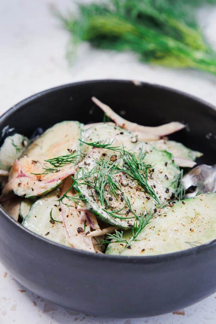 A black bowl filled with a cucumber and onion salad, garnished with fresh dill and cracked black pepper. A fork rests inside the bowl, and fresh dill is blurred in the background.