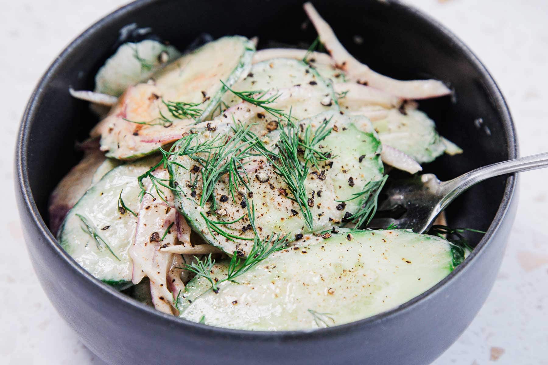 A black bowl filled with cucumber salad, thinly sliced cucumbers and onions, topped with fresh dill and cracked black pepper, with a metal fork inside the bowl.