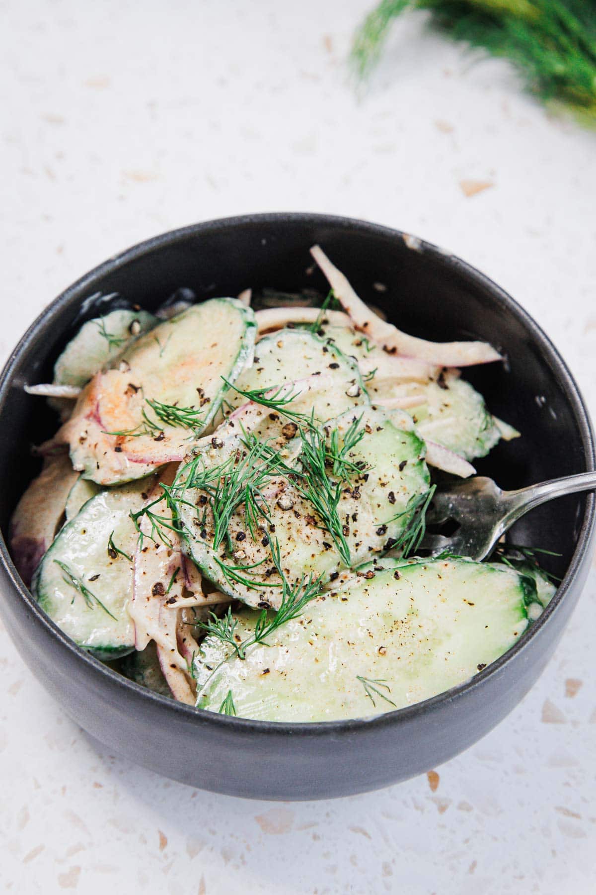 A black bowl filled with cucumber salad, featuring sliced cucumbers, onions, a creamy dressing, fresh dill, and cracked black pepper. A fork rests inside the bowl on a light, speckled surface.