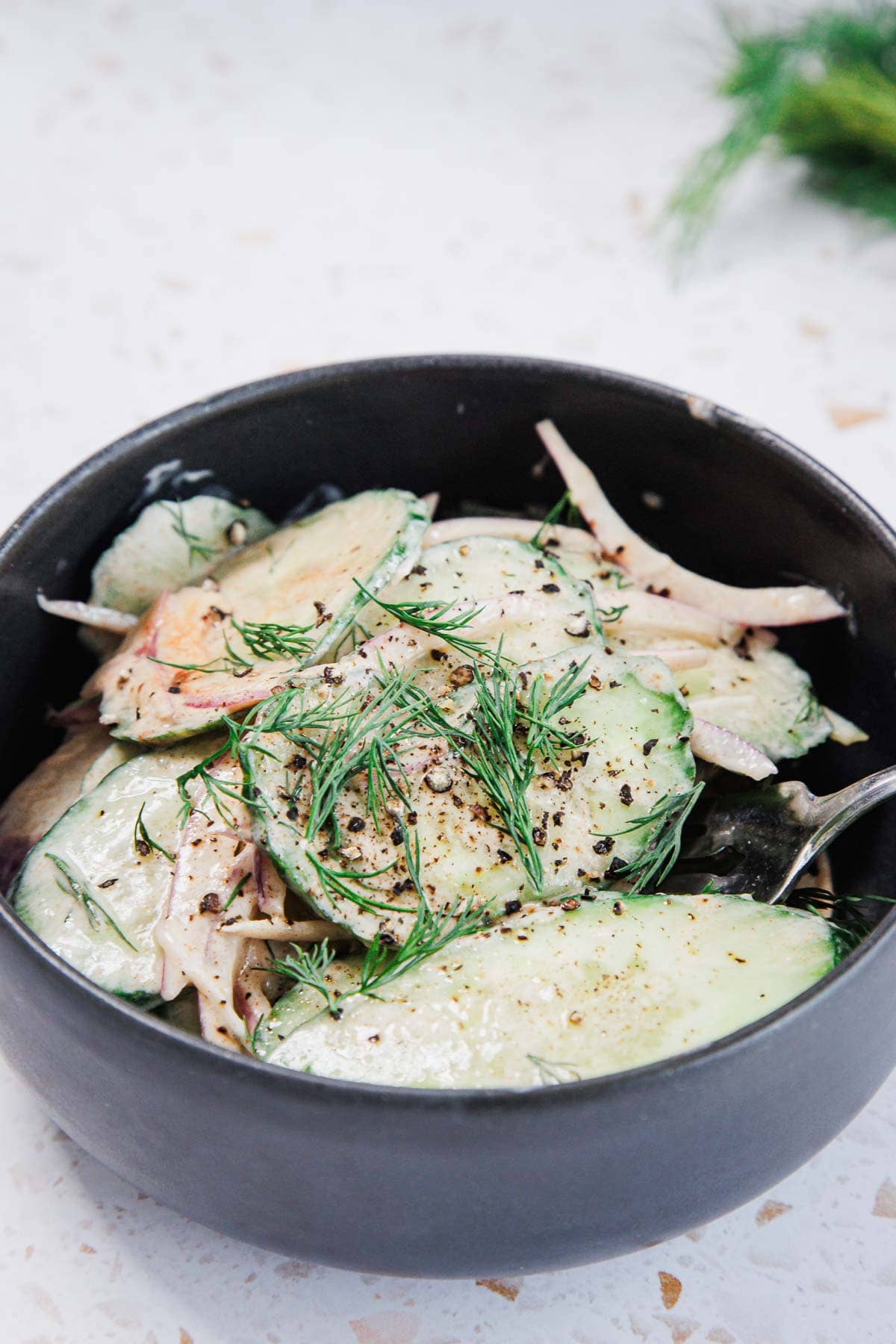 A black bowl filled with a salad of sliced cucumbers, thin onion strips, fresh dill, and cracked black pepper, with a fork partially visible inside the bowl.