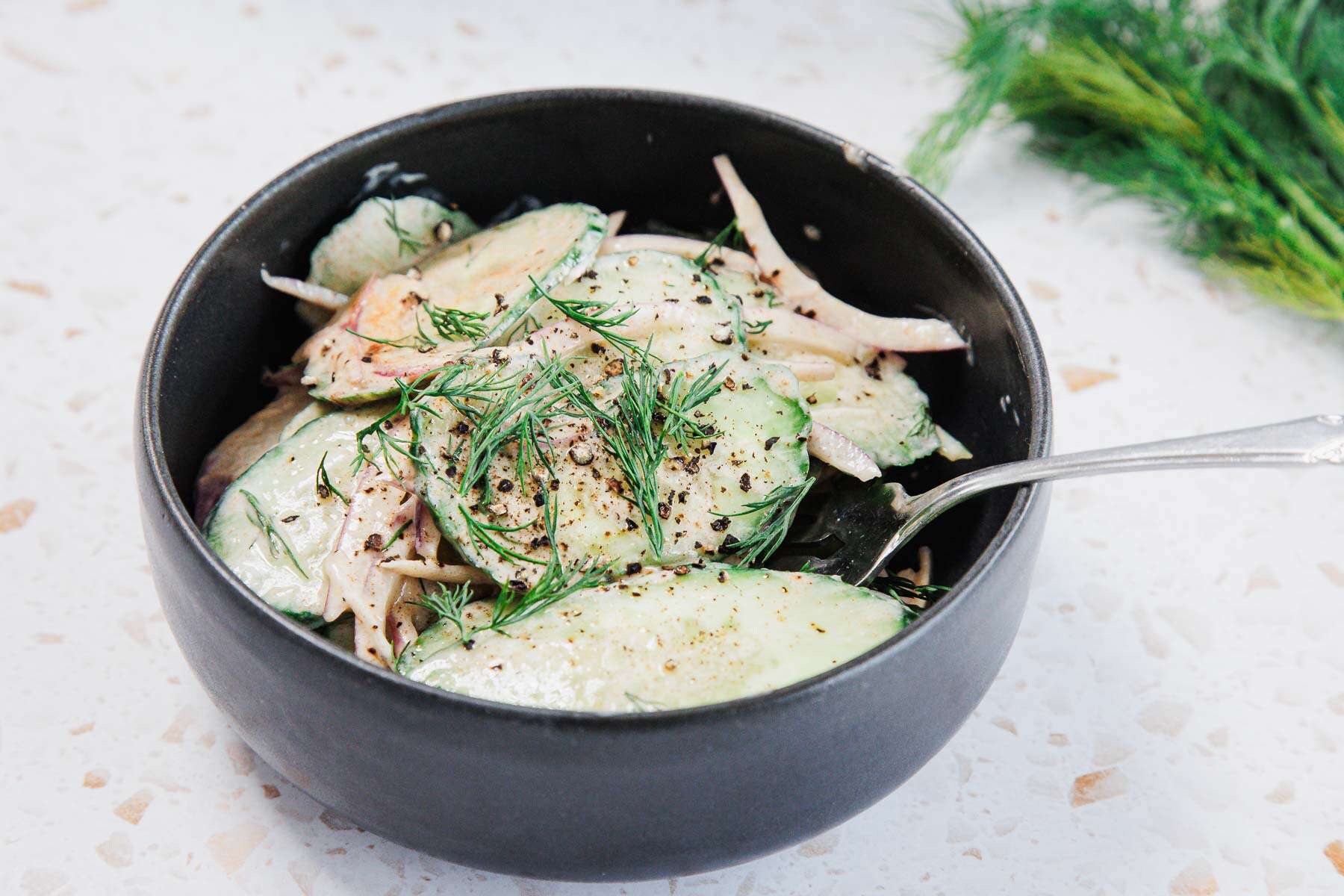 A black bowl filled with sliced cucumbers, onions, and fresh dill, topped with cracked black pepper and a creamy dressing. A spoon rests in the bowl and dill garnish is on the table in the background.