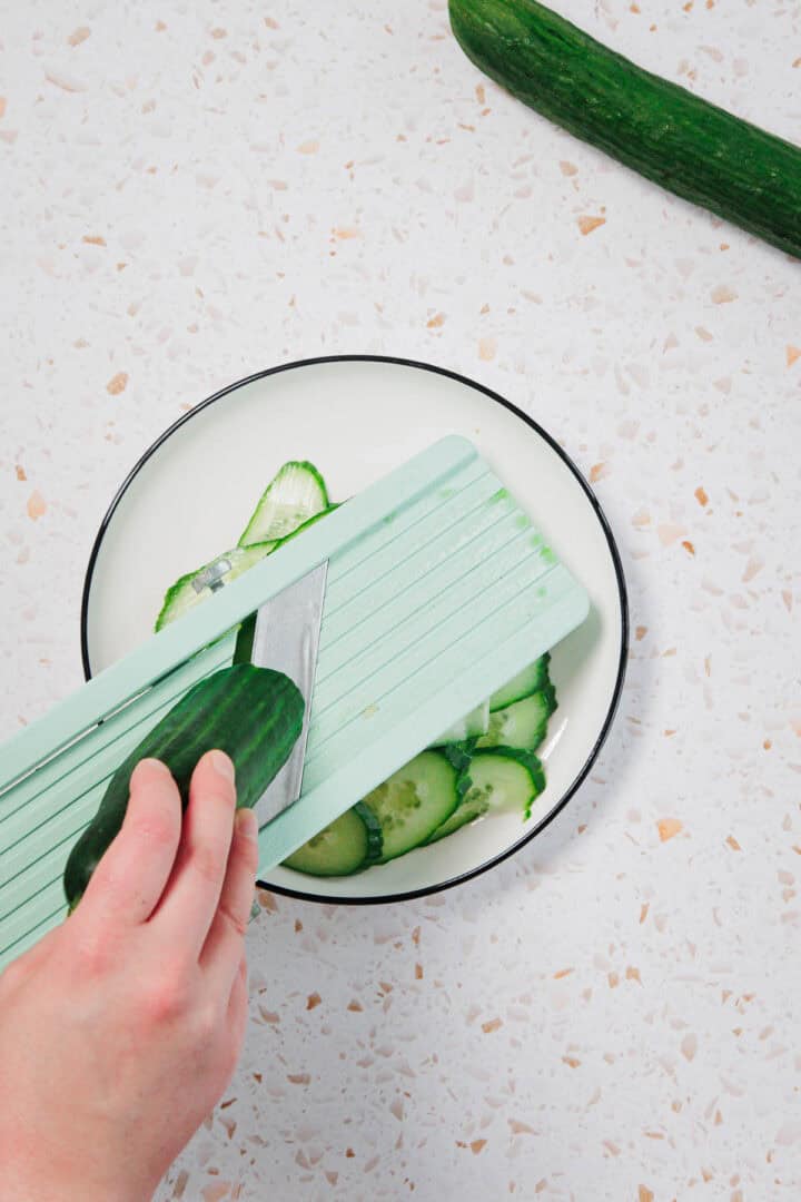 A hand slices a cucumber with a light green mandoline slicer over a white plate containing several thin cucumber slices. Another whole cucumber is visible on the countertop.