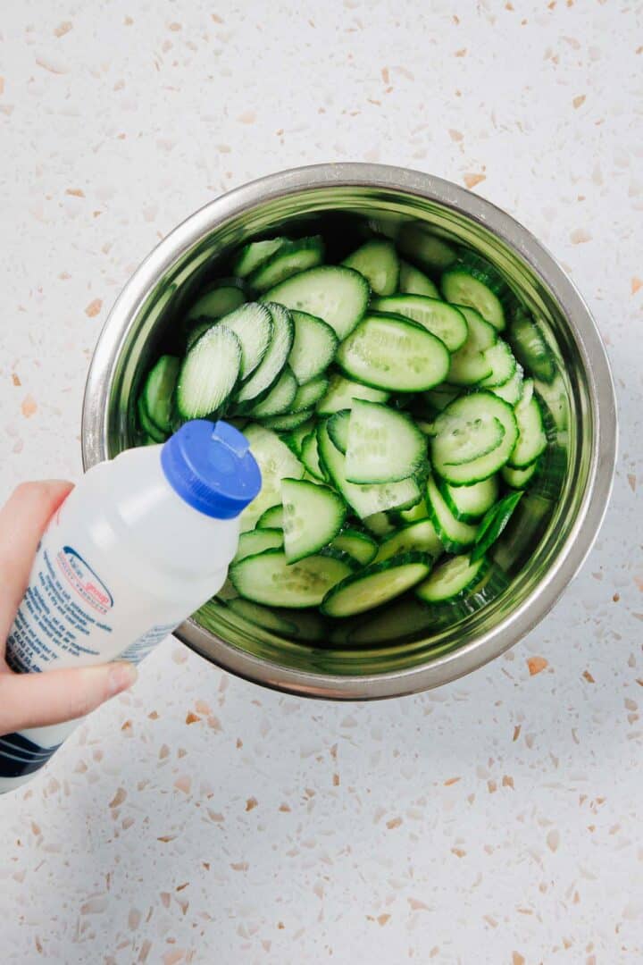A hand holding a bottle of liquid above a metal bowl filled with sliced cucumbers, positioned on a light-colored, speckled countertop.