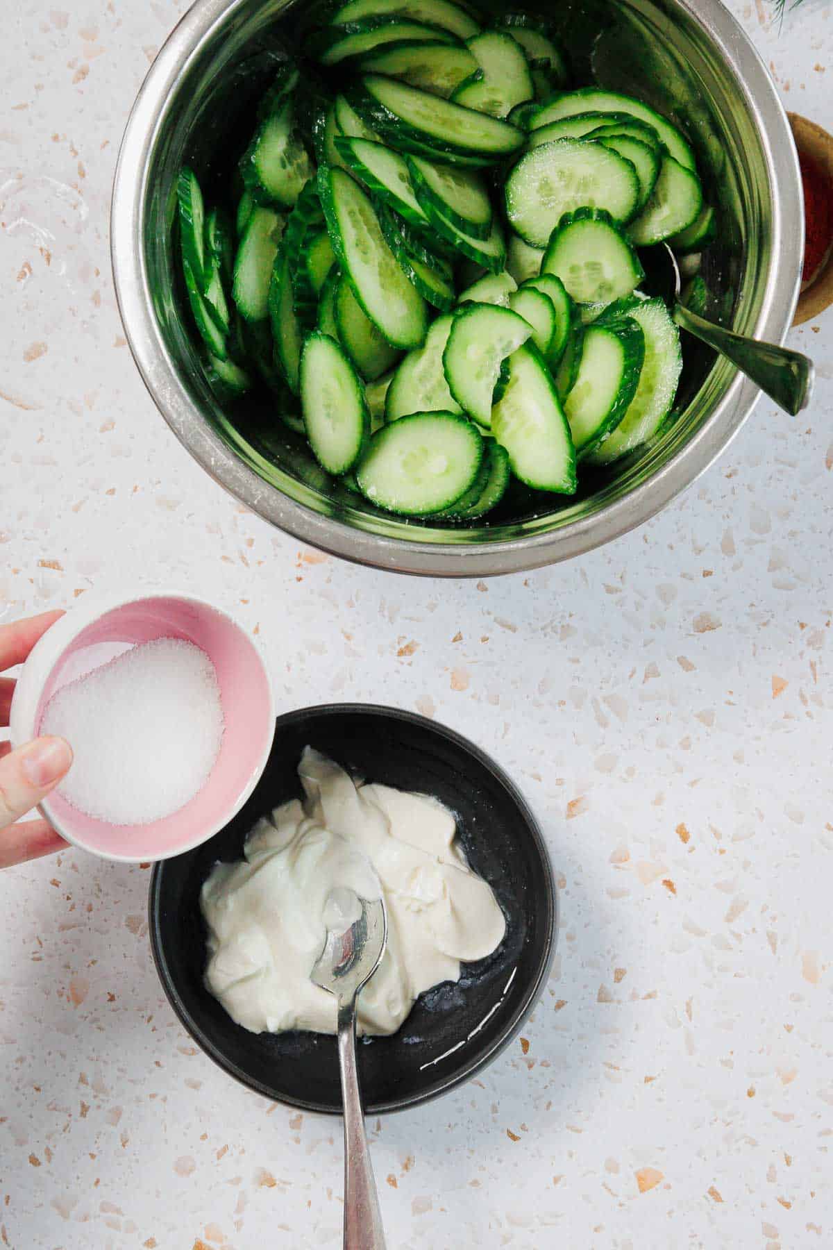 A metal bowl of sliced cucumbers with a spoon, a small black bowl of mayonnaise or yogurt with a spoon, and a hand holding a bowl of sugar above the black bowl, all on a light-colored counter.