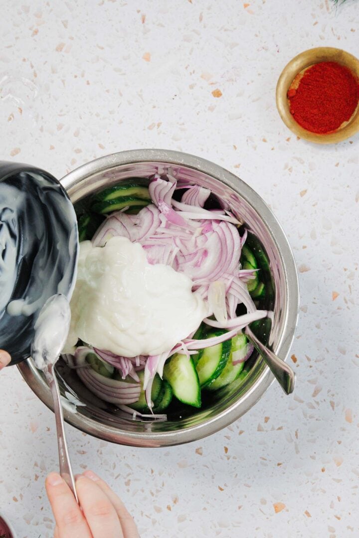 A person pours yogurt from a black bowl into a metal mixing bowl containing sliced red onions and cucumbers. A bowl of red spice sits nearby on a white countertop.
