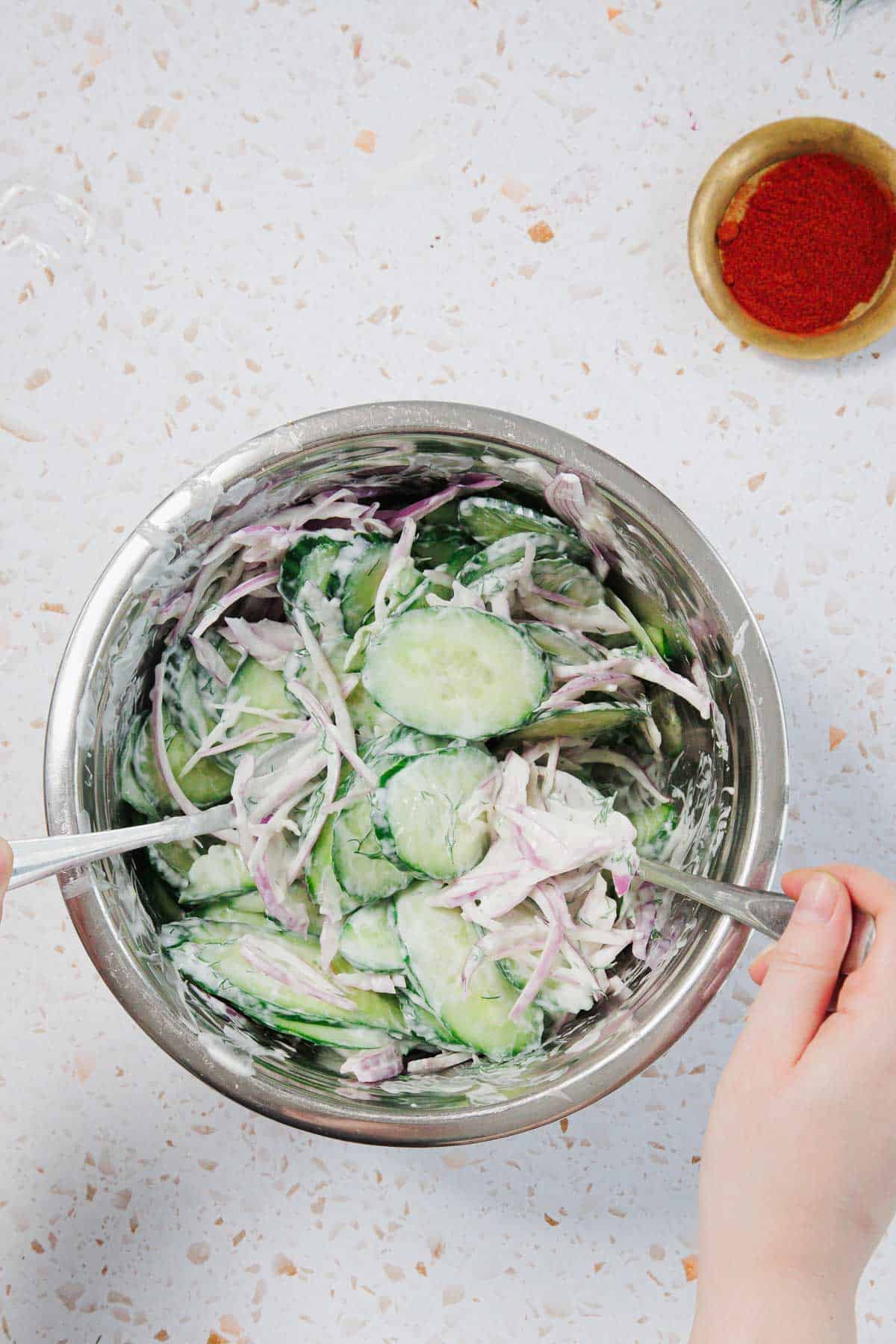 A person mixes sliced cucumbers and red onions coated in a creamy dressing in a stainless steel bowl with two forks. A small bowl of paprika sits nearby on a white speckled countertop.