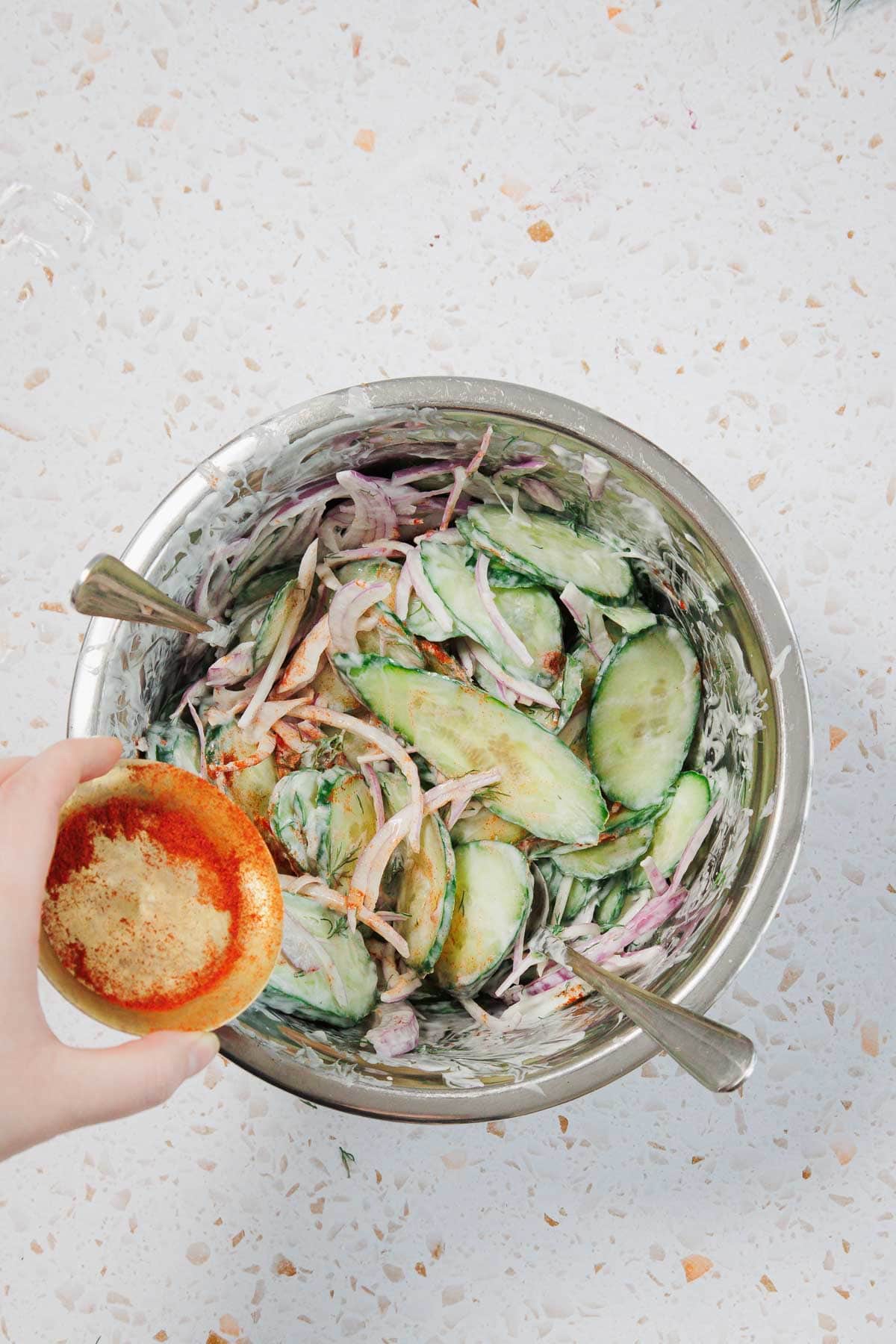 A hand holds a halved grapefruit topped with brown sugar above a metal bowl containing a salad of sliced cucumbers, onions, and creamy dressing. Two utensils are in the bowl on a white speckled surface.