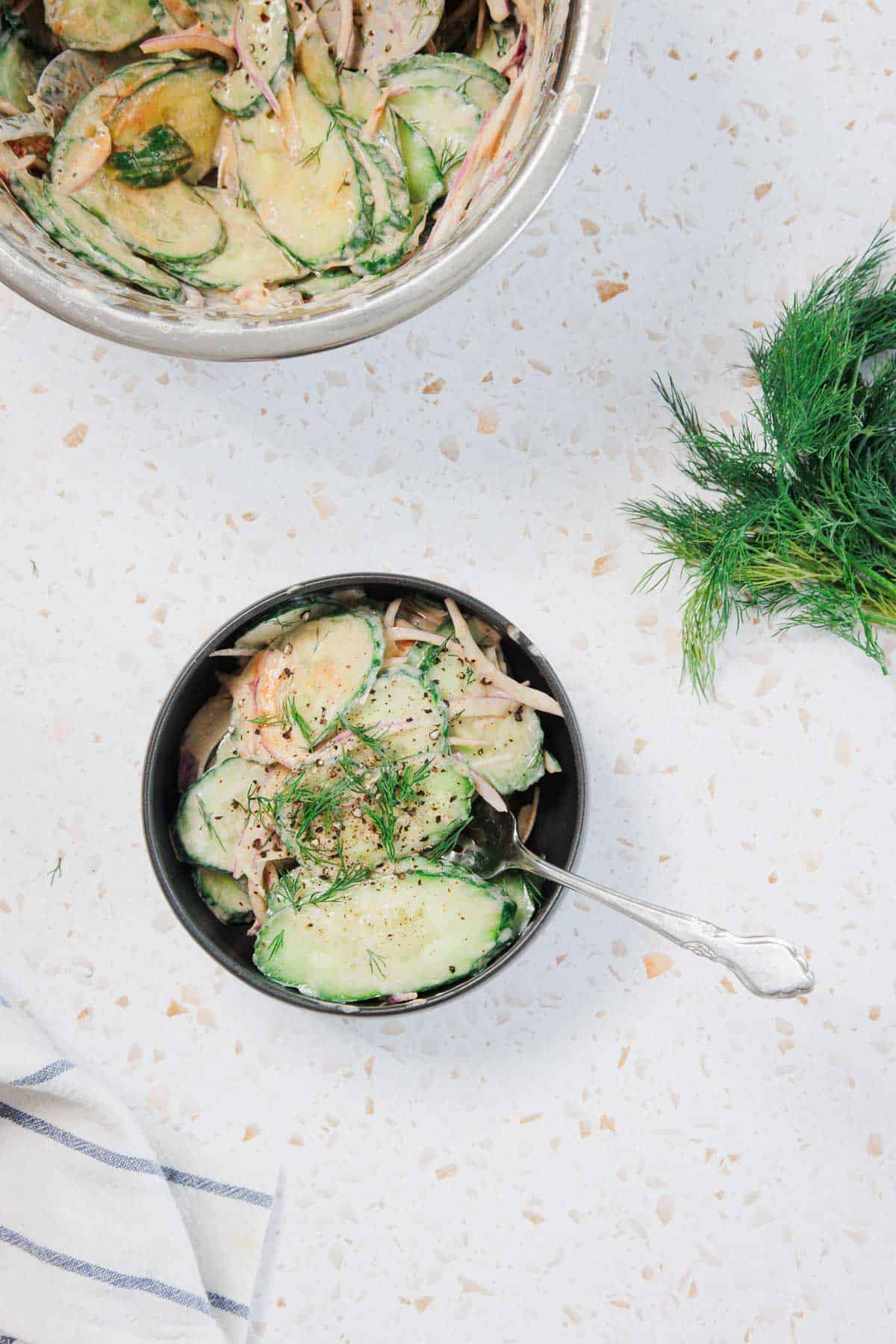 A bowl of creamy cucumber salad with dill, placed on a white countertop next to a larger mixing bowl of salad and a bunch of fresh dill, with a striped cloth partially visible in the corner.