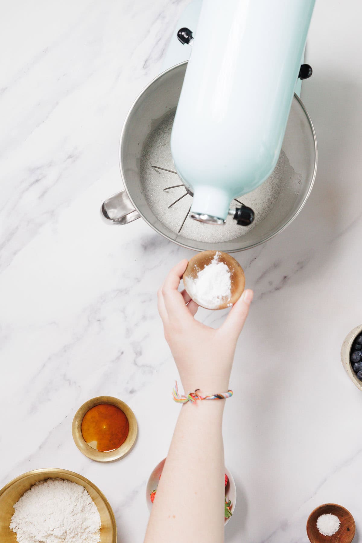 A hand holds a small wooden bowl of white powder over a stand mixer with flour inside, prepping for Gluten-Free Angel Food Cake. Nearby are bowls of liquid, flour, salt, and blueberries on a white marble surface.