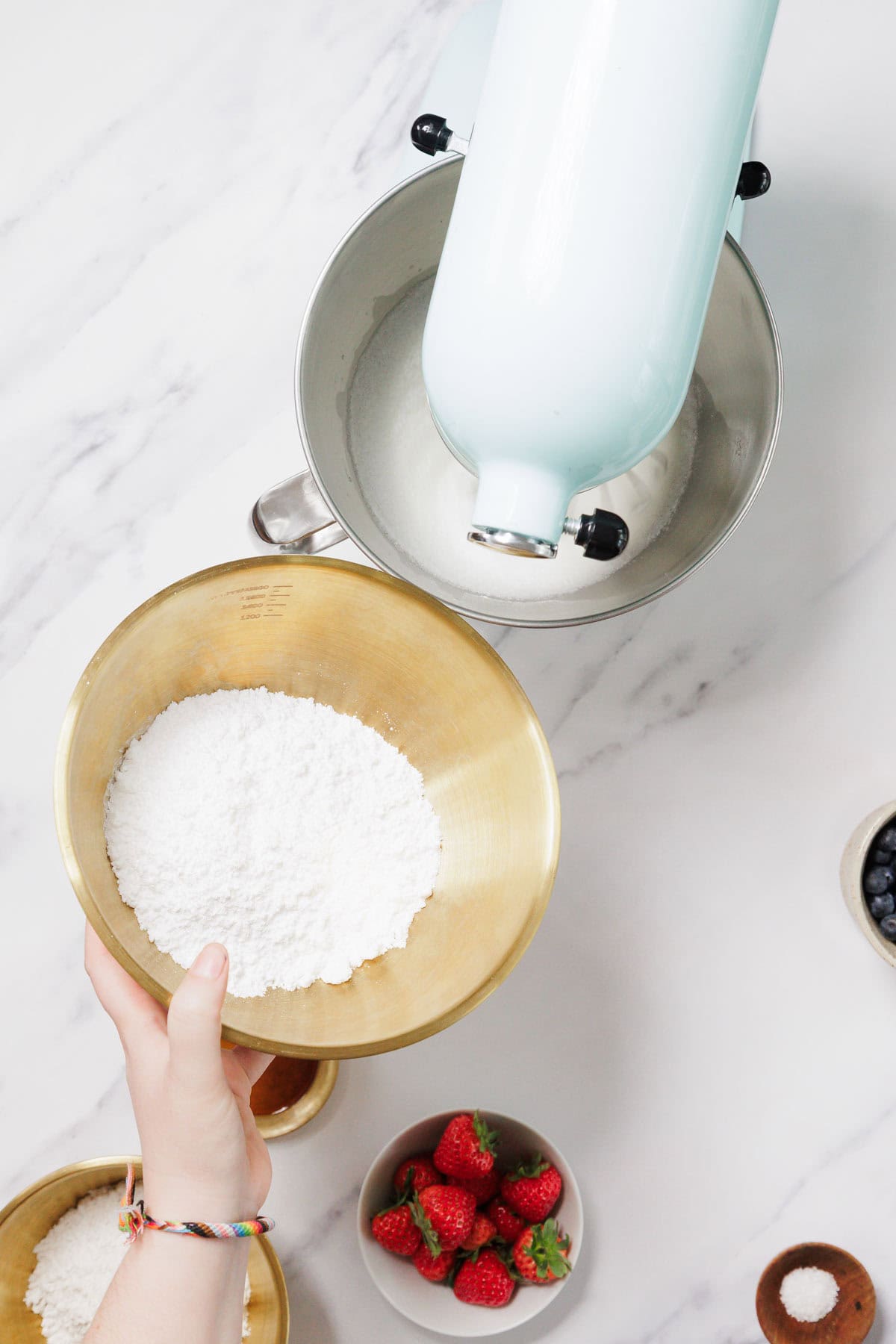 A hand holds a bowl of powdered sugar above a stand mixer with whipped ingredients for Gluten-Free Angel Food Cake. Fresh strawberries and blueberries are in small bowls on the white countertop nearby.