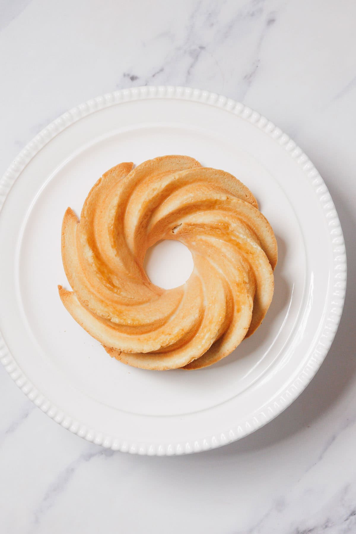 A golden brown Gluten-Free Angel Food Cake with a swirl pattern sits on a white, round plate with a beaded edge, placed on a white marble surface.