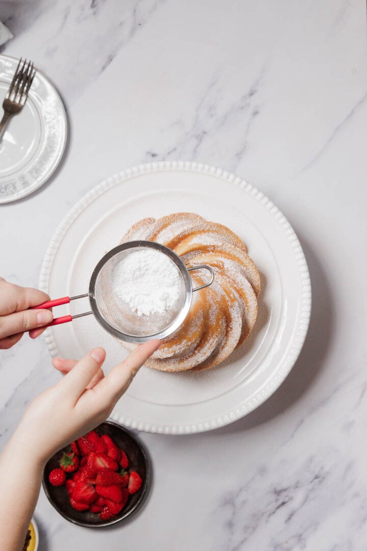 A person sifts powdered sugar onto a Gluten-Free Angel Food Cake on a white plate. Sliced strawberries in a bowl and a fork on a small plate are nearby on the marble surface.