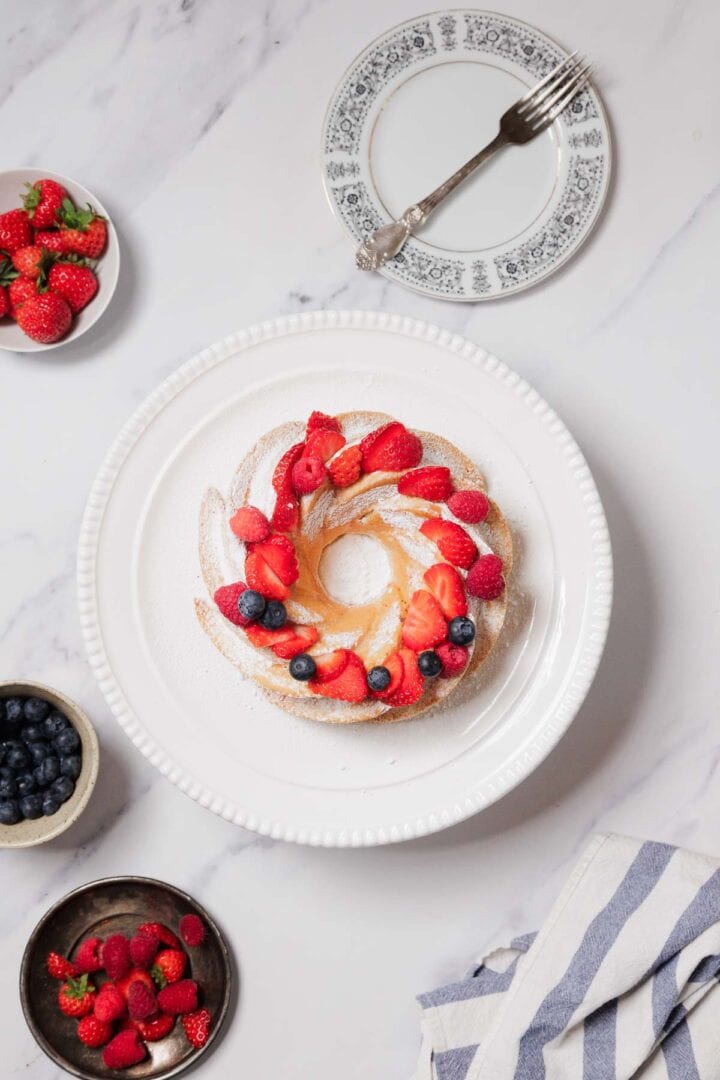 A Gluten-Free Angel Food Cake topped with powdered sugar, strawberries, raspberries, and blueberries sits on a white cake stand. Nearby are plates of berries, an empty plate with a fork, and a striped towel on a marble surface.