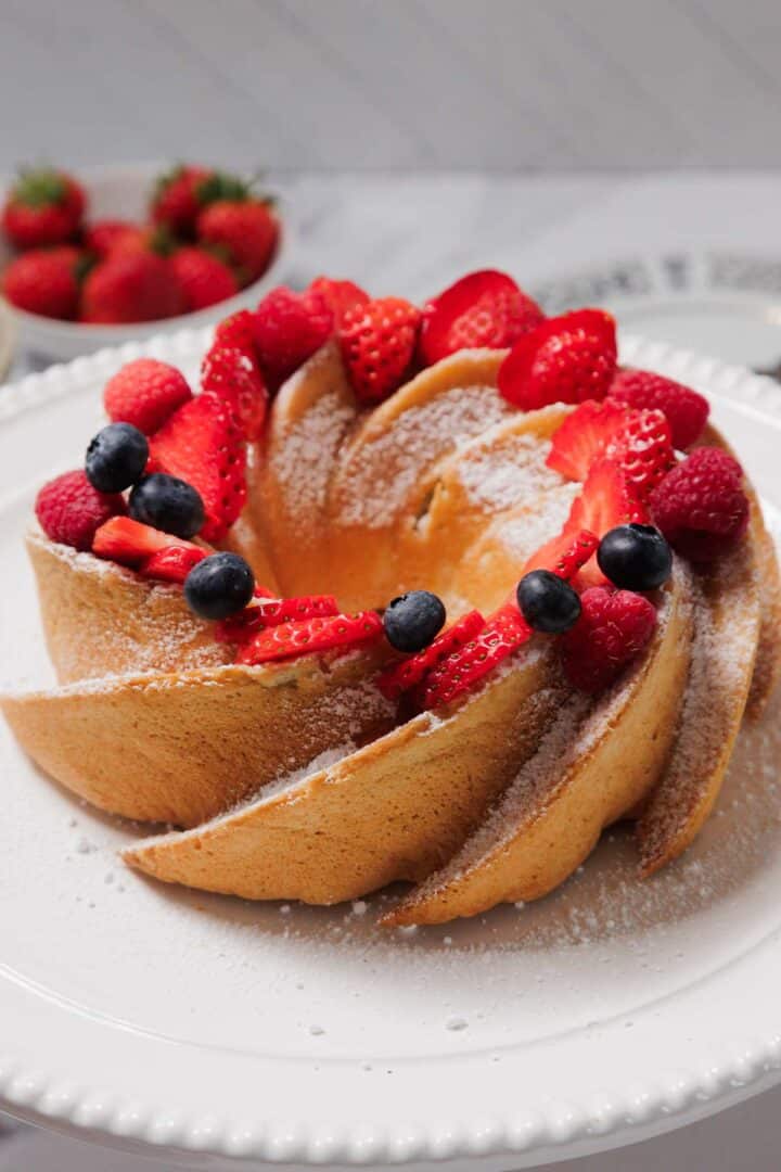 A Gluten-Free Angel Food Cake dusted with powdered sugar sits on a white plate, decorated with fresh sliced strawberries, raspberries, and blueberries. A bowl of whole strawberries is in the background.