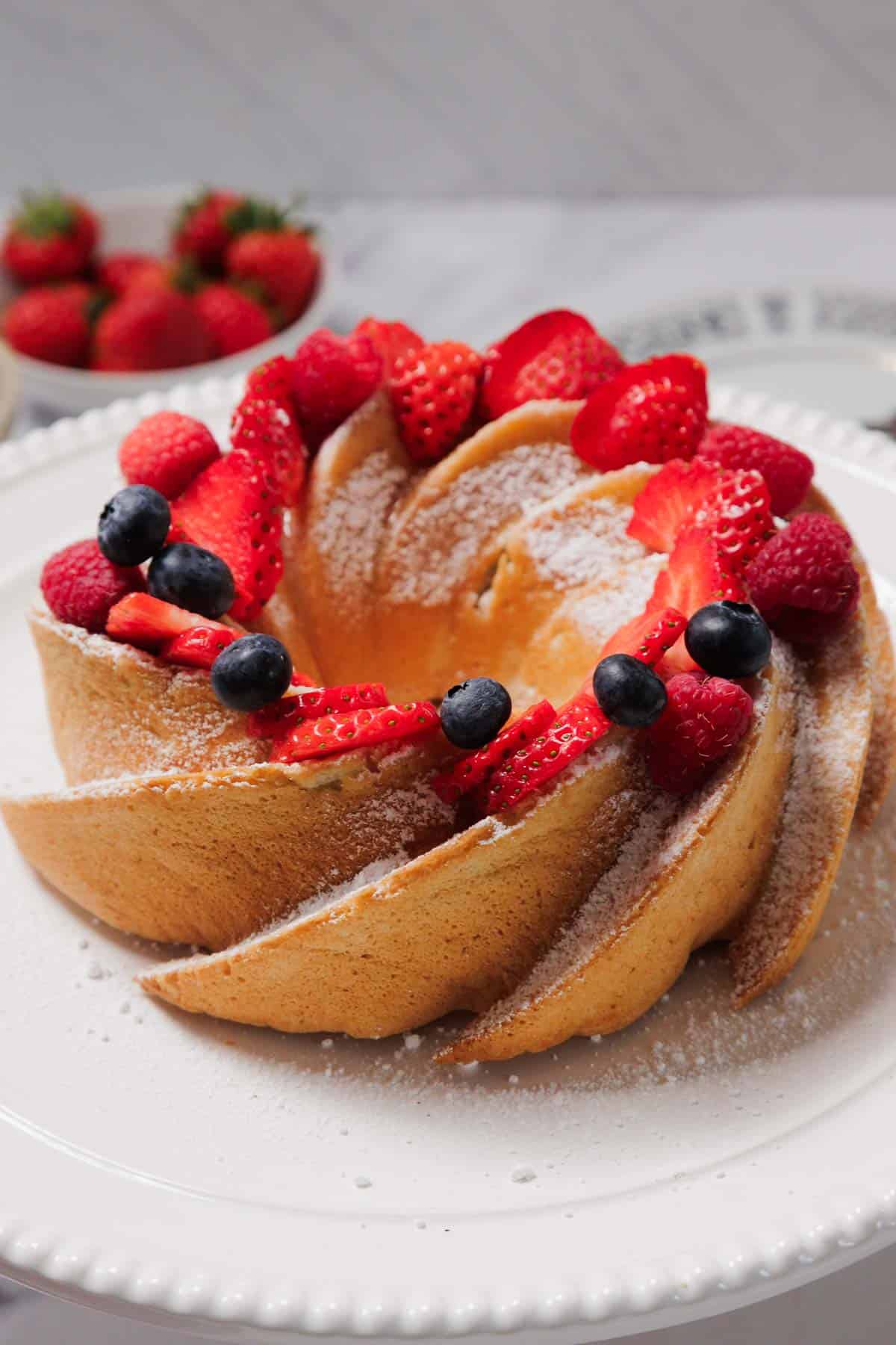 A Gluten-Free Angel Food Cake dusted with powdered sugar sits on a white plate, decorated with fresh sliced strawberries, raspberries, and blueberries. A bowl of whole strawberries is in the background.