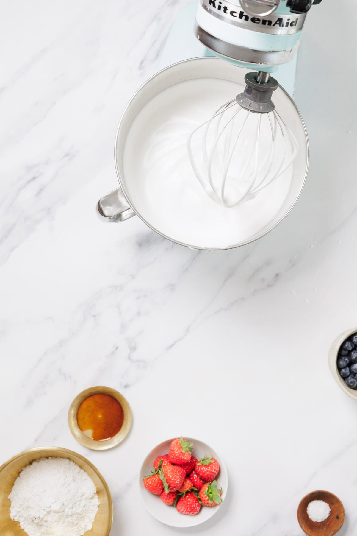 A stand mixer with a whisk attachment is mixing whipped cream in a metal bowl. Nearby are small bowls containing flour for Gluten-Free Angel Food Cake, strawberries, blueberries, and a liquid ingredient on a white marble surface.