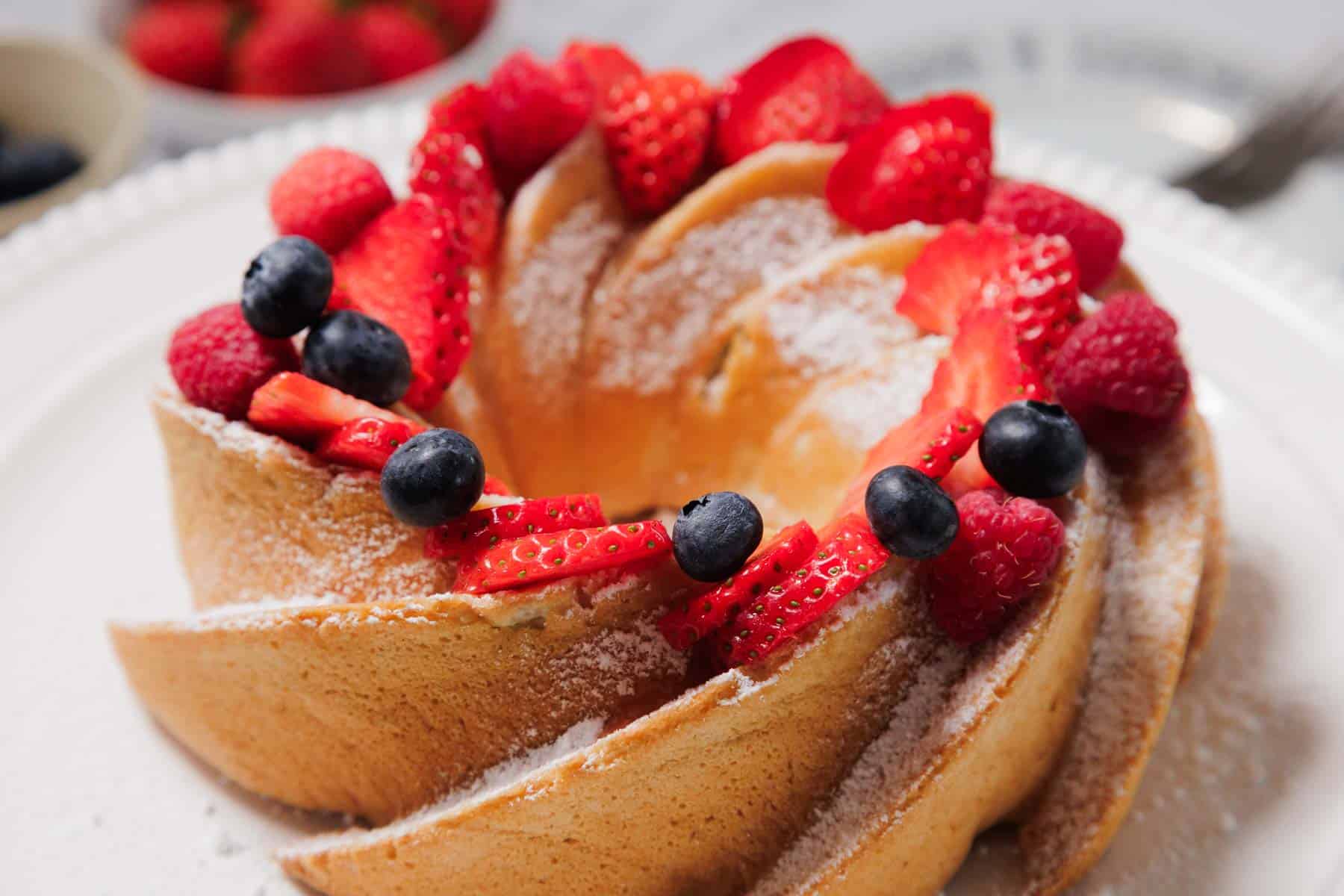 A Gluten-Free Angel Food Cake topped with powdered sugar, fresh strawberries, raspberries, and blueberries sits on a white plate.