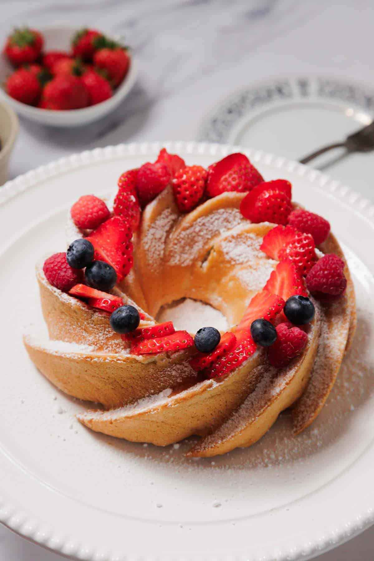 A Gluten-Free Angel Food Cake dusted with powdered sugar is decorated with sliced strawberries and whole blueberries. The cake sits on a white plate, with a bowl of strawberries and two empty plates in the background.