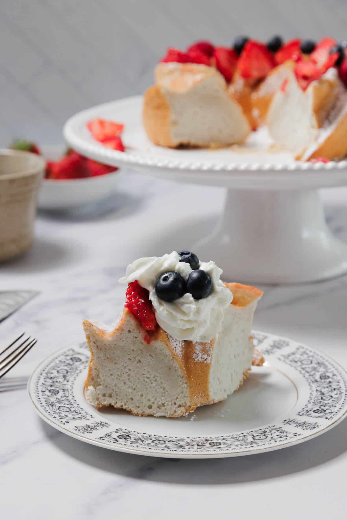 A slice of Gluten-Free Angel Food Cake on a patterned plate, topped with whipped cream, blueberries, and a strawberry. In the background, the remaining cake topped with berries rests on a white cake stand.