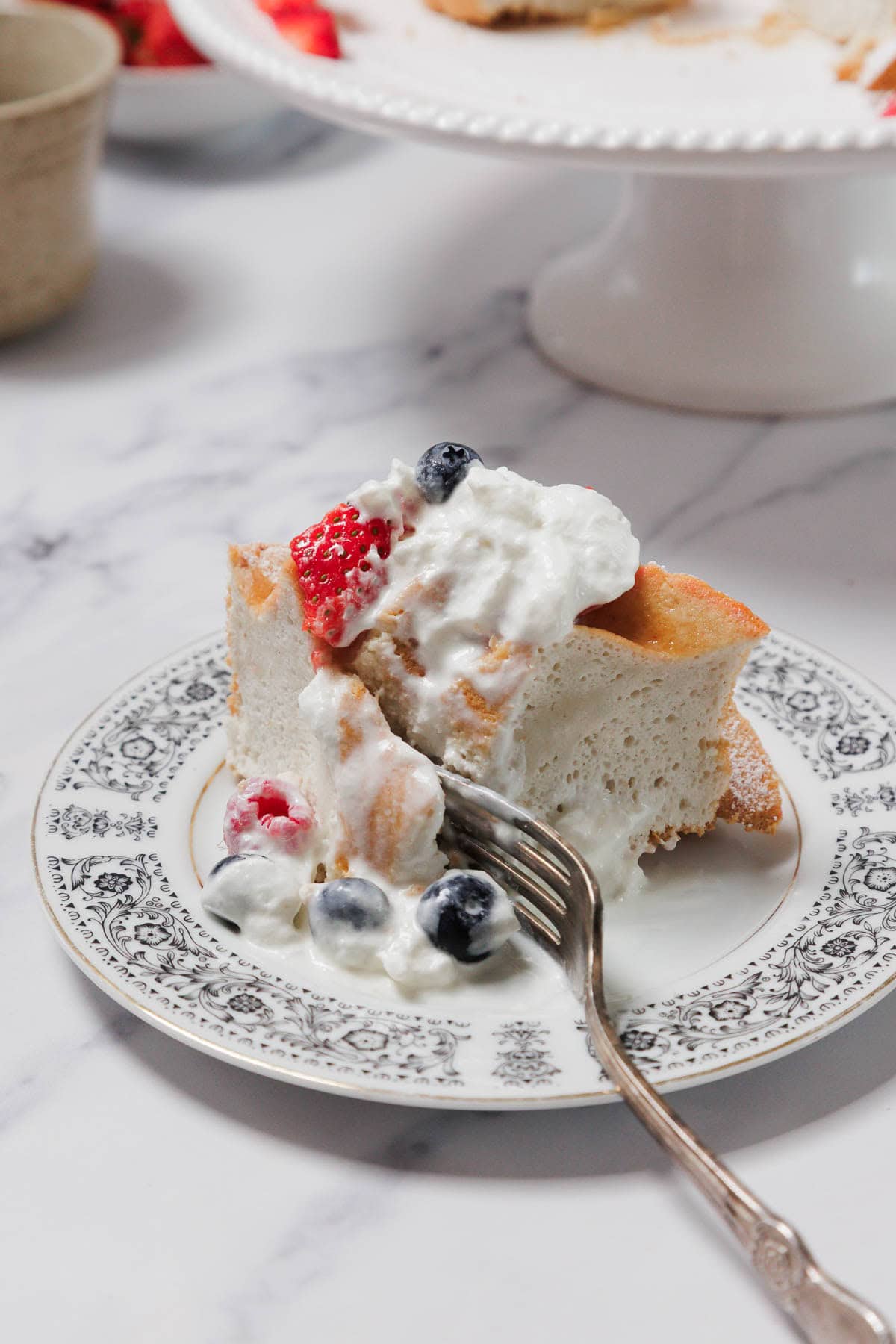 A slice of Gluten-Free Angel Food Cake topped with whipped cream and berries sits on a decorative plate. A fork rests on the plate, with some cake and cream on its tines. A cake stand and bowls are blurred in the background.