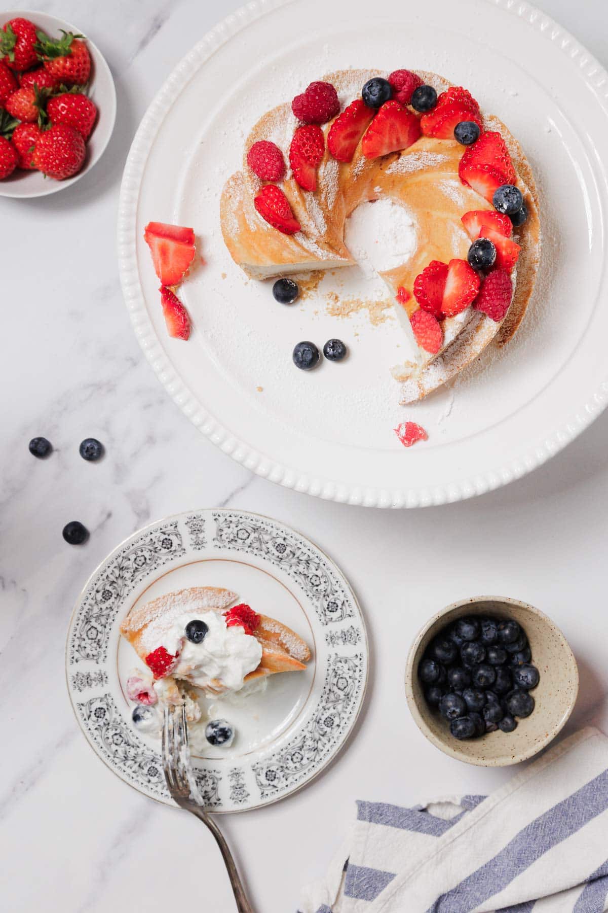 A partly sliced Gluten-Free Angel Food Cake topped with strawberries, raspberries, and blueberries sits on a white plate. A serving of the cake is on a smaller patterned plate with a fork, accompanied by bowls of strawberries and blueberries.