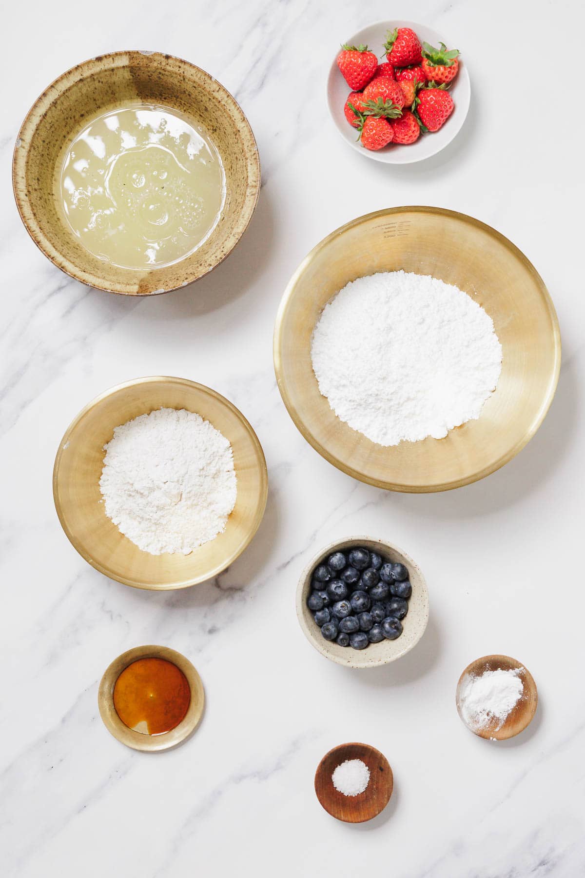 Various baking ingredients arranged on a white marble surface, including bowls of flour for Gluten-Free Angel Food Cake, powdered sugar, egg white, blueberries, a small plate of strawberries, honey, and small dishes with salt and baking powder.