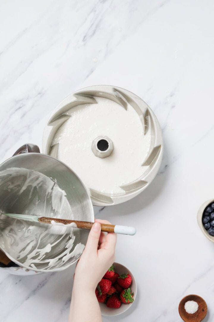A person pours Gluten-Free Angel Food Cake batter from a metal mixing bowl into a bundt cake pan on a white marble surface. Nearby are bowls of strawberries, blueberries, and a small dish of salt.