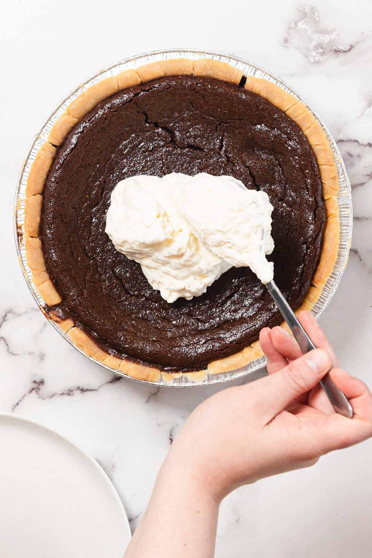 A hand is spreading a large dollop of whipped cream onto a baked chocolate pie in a foil pie dish on a marble countertop. A white plate is partially visible in the lower left corner.