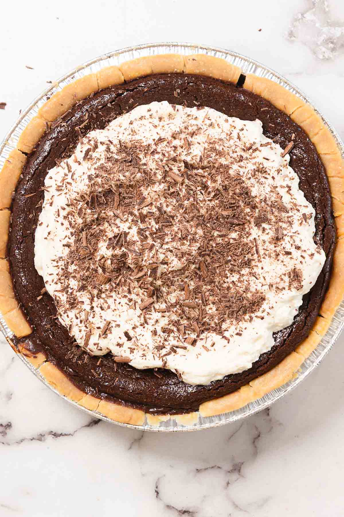 A chocolate pie in a foil pie pan with a baked crust, topped with whipped cream and chocolate shavings, placed on a marble countertop.