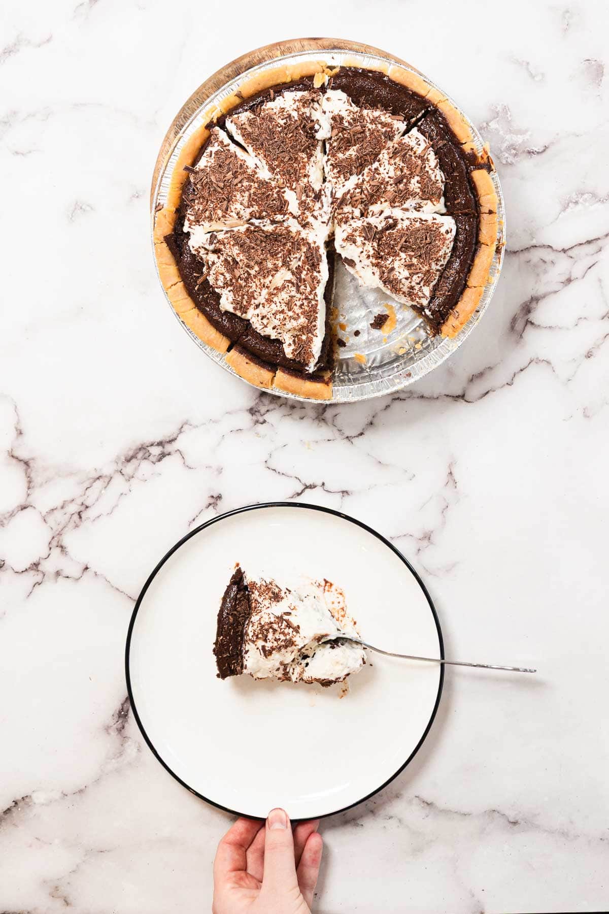 A hand holds a plate with a slice of chocolate cream pie topped with whipped cream and chocolate shavings. The rest of the pie, with several pieces missing, sits above on a marble surface.