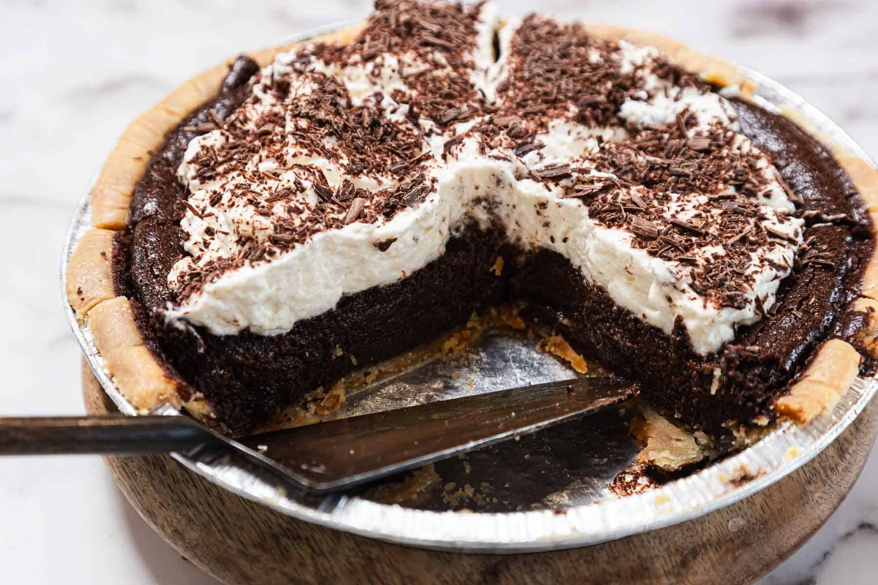 A chocolate pie topped with whipped cream and chocolate shavings in a metal pie tin, with a slice missing and a pie server resting inside the tin.