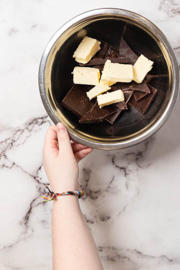A hand holds a metal bowl filled with chunks of dark chocolate and pieces of butter, set on a white marble surface. The person is wearing a colorful braided bracelet.