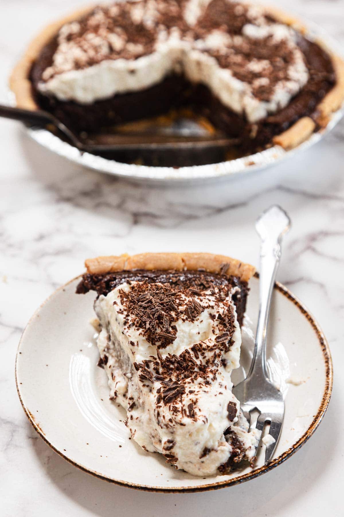 A slice of chocolate cream pie topped with whipped cream and chocolate shavings sits on a plate with a fork. The rest of the pie is in the background with one slice missing.