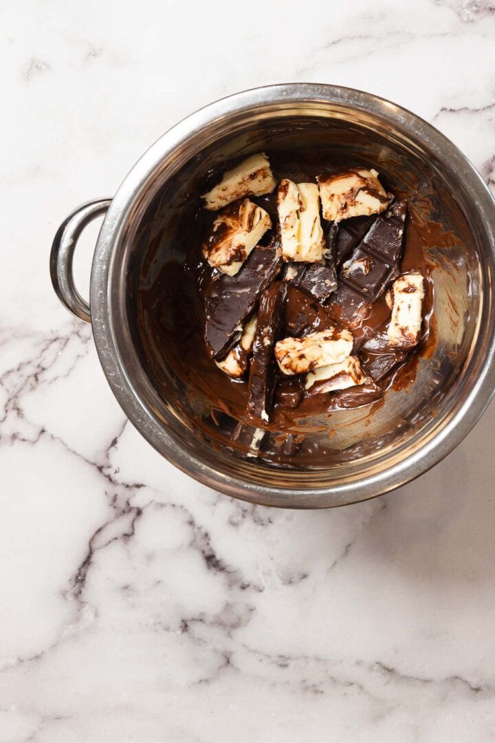 A stainless steel bowl containing pieces of dark chocolate and chunks of butter, partially melted, sits on a white marble countertop.
