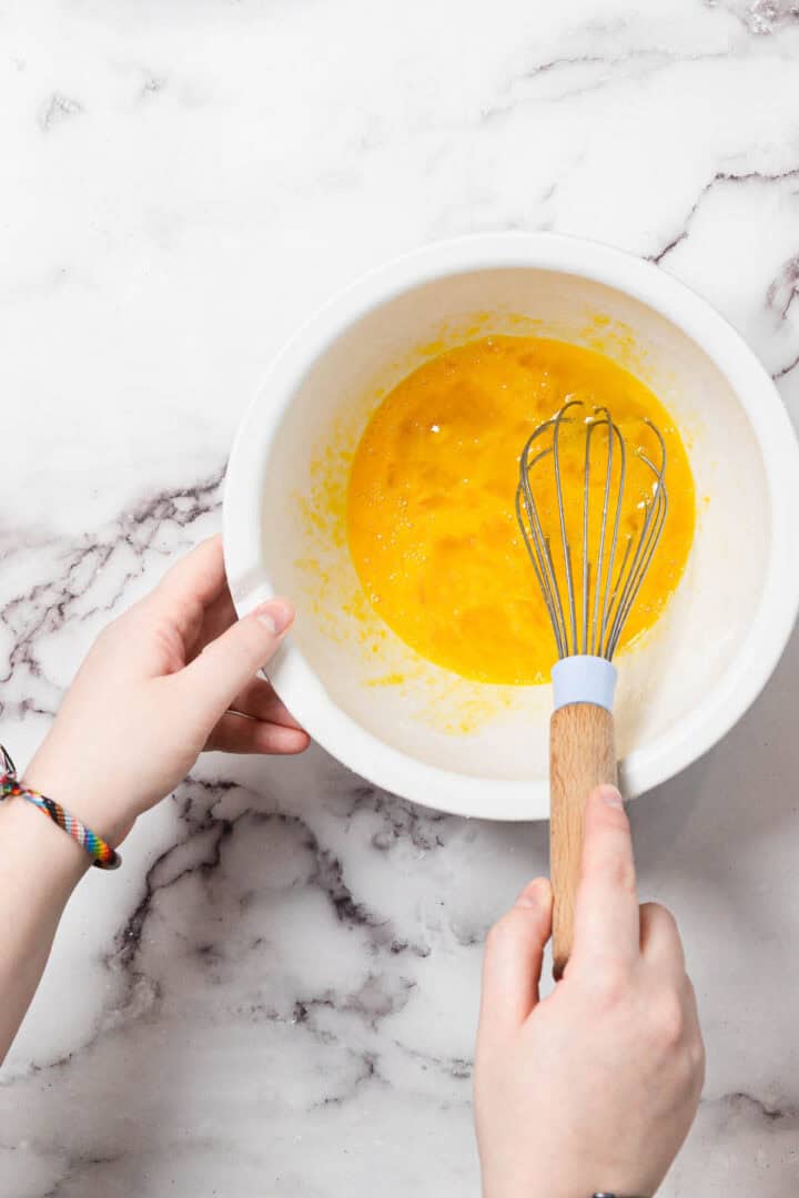 A person holds a white bowl with beaten eggs in it while whisking with a metal whisk on a white marble surface.