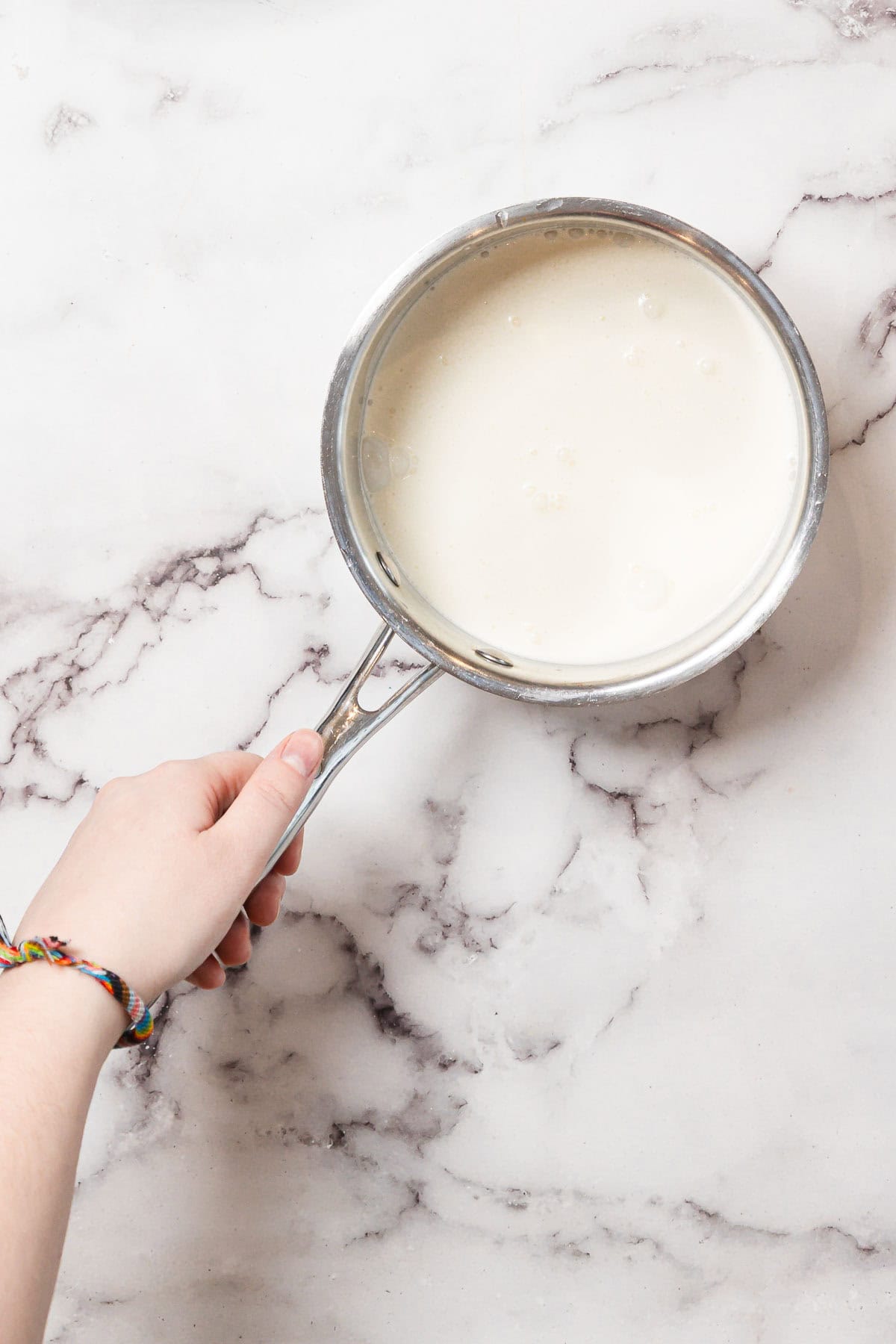 A hand holding a metal saucepan filled with a white liquid, likely milk, over a white marble countertop. The person is wearing a multicolored braided bracelet on their wrist.